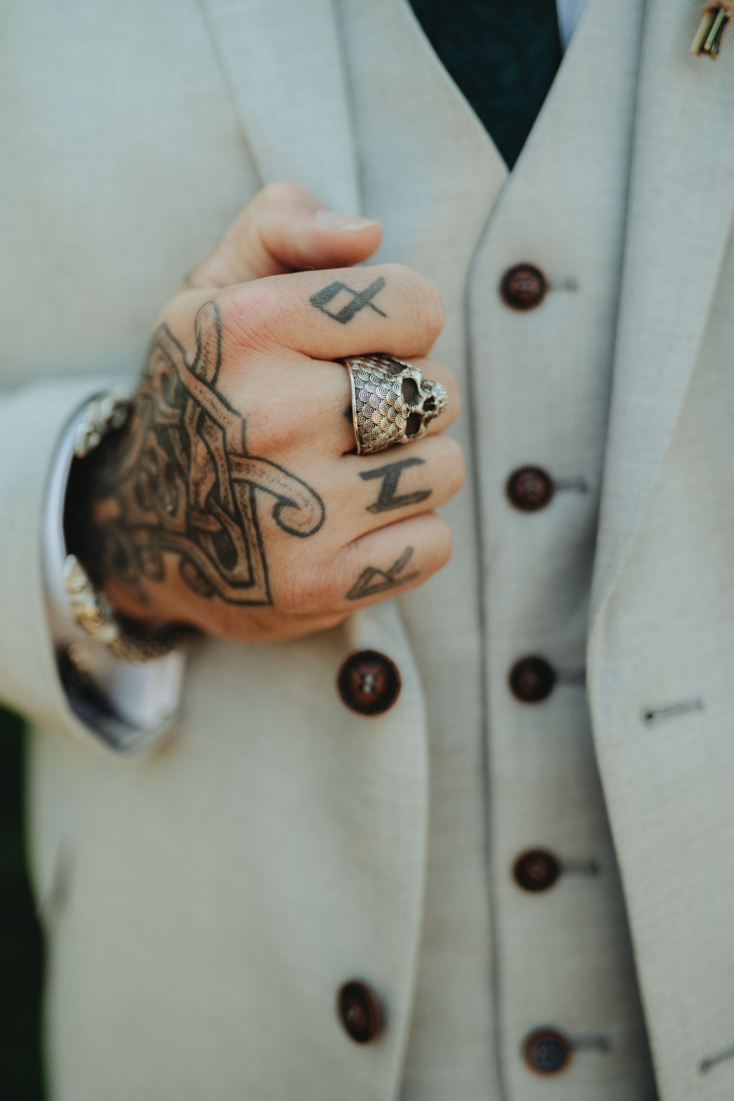 Tattooed hand with skull ring in a light gray suit, showcasing bold ink designs and ornate jewelry.