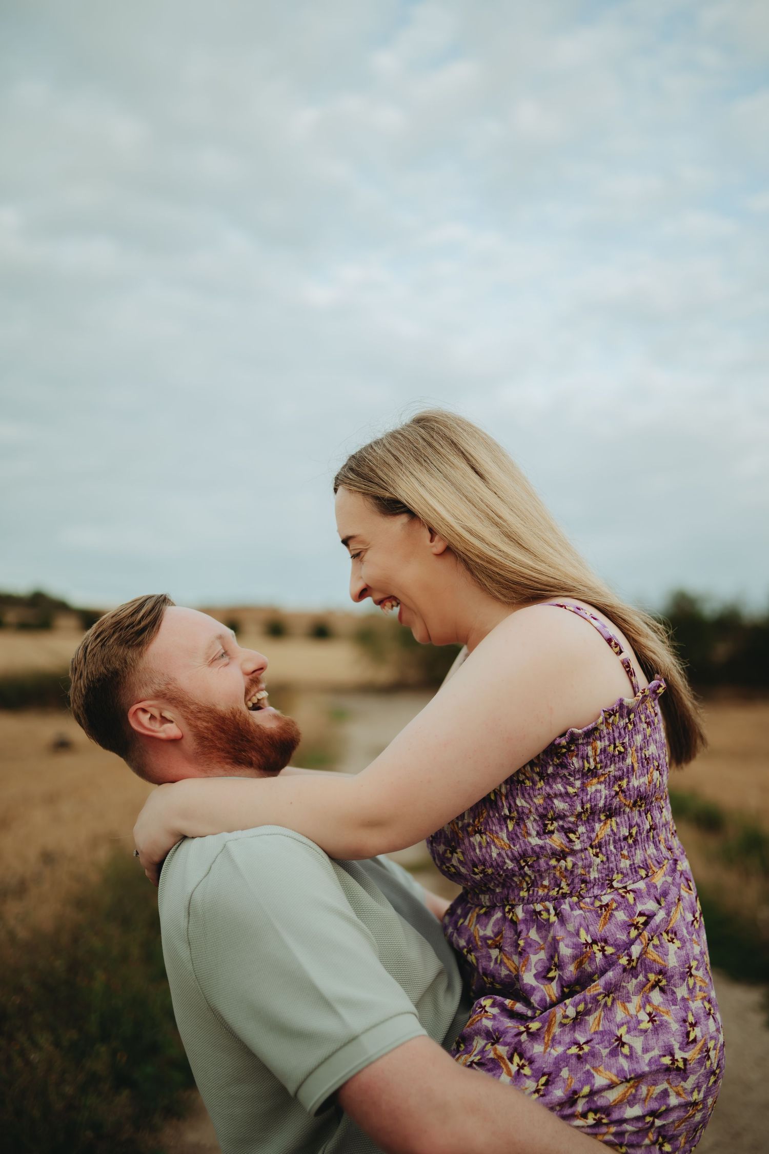 A joyful couple laughs together outdoors, the man lifting the woman in a purple floral dress against a golden field backdrop.