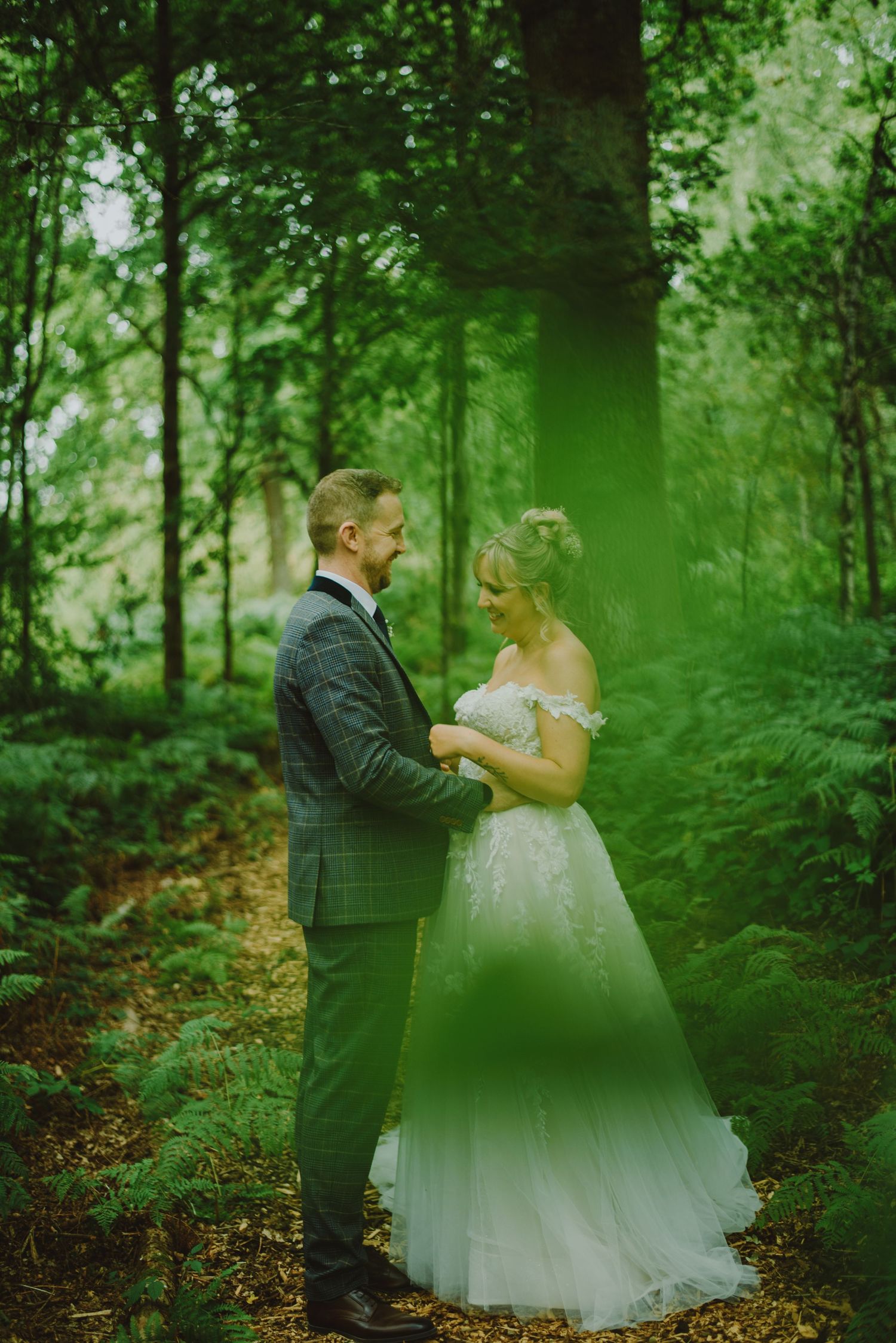 A couple in wedding attire embraces in a lush green forest setting during their special day.