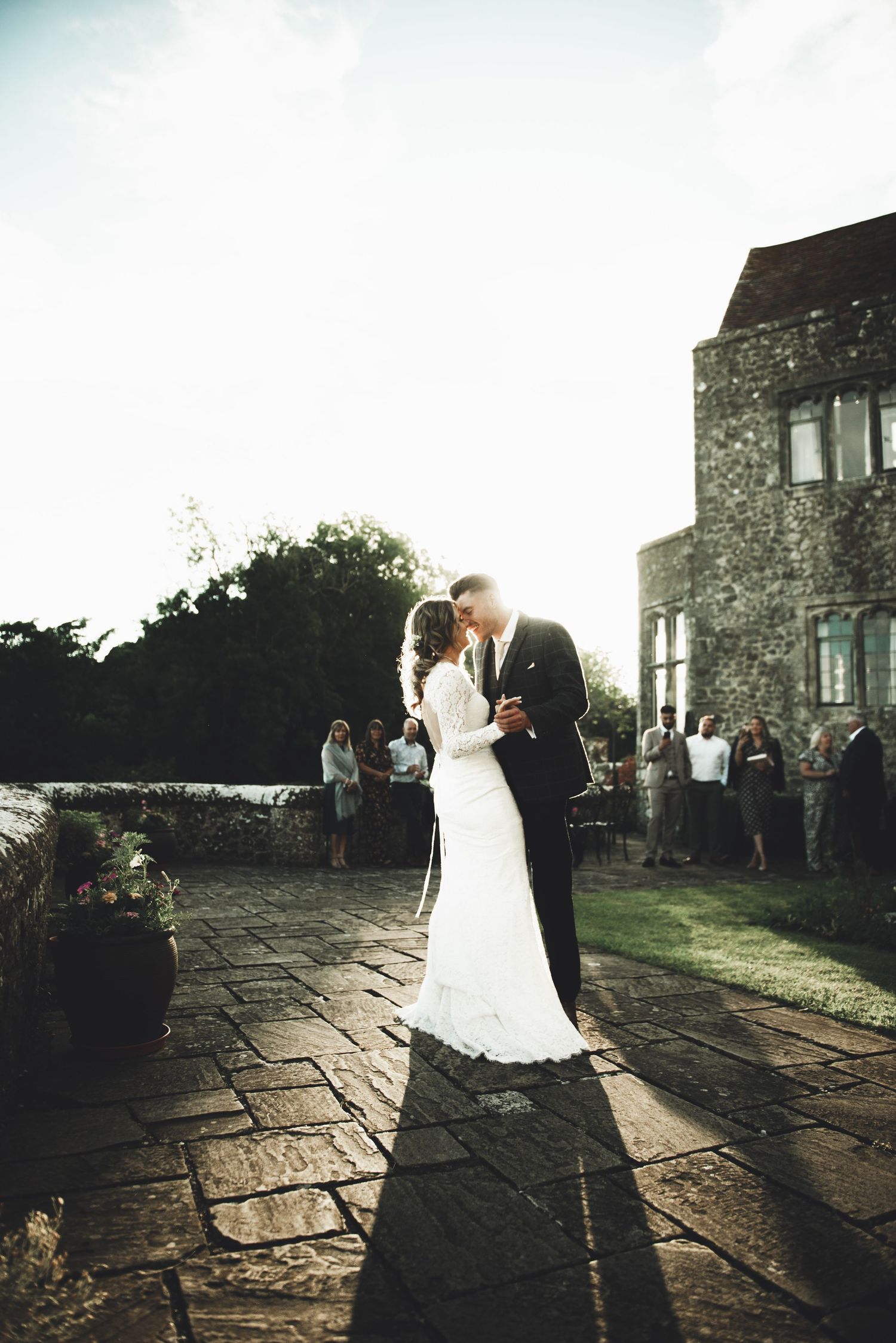 Wedding couple silhouetted against dramatic sunlight on stone patio during outdoor castle ceremony.