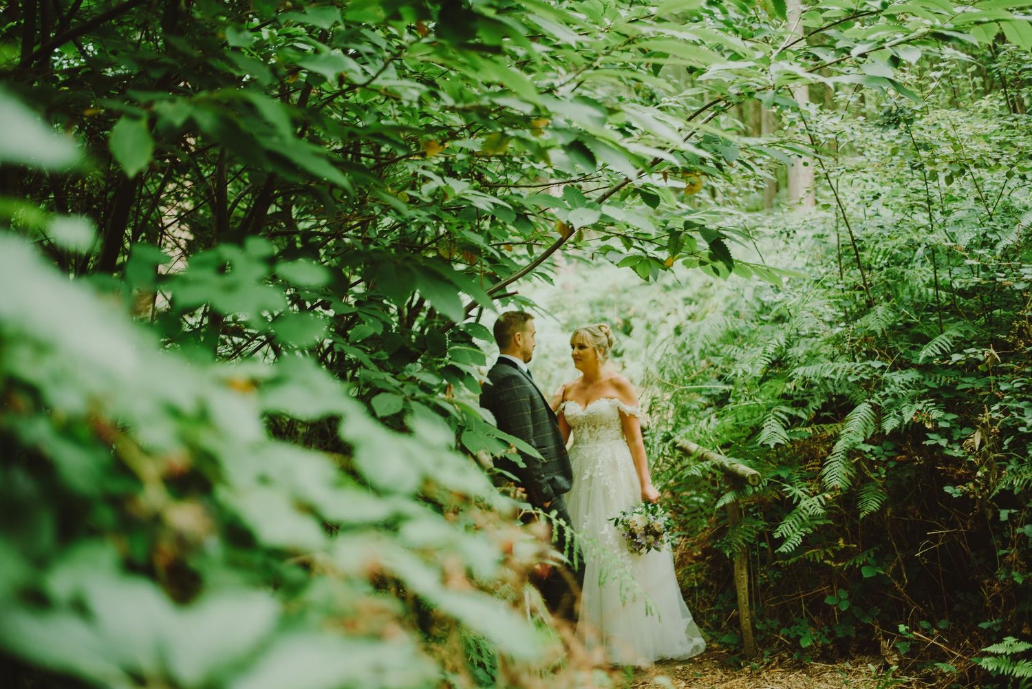 A couple stands together on a lush forest path surrounded by vibrant green foliage on their wedding day.