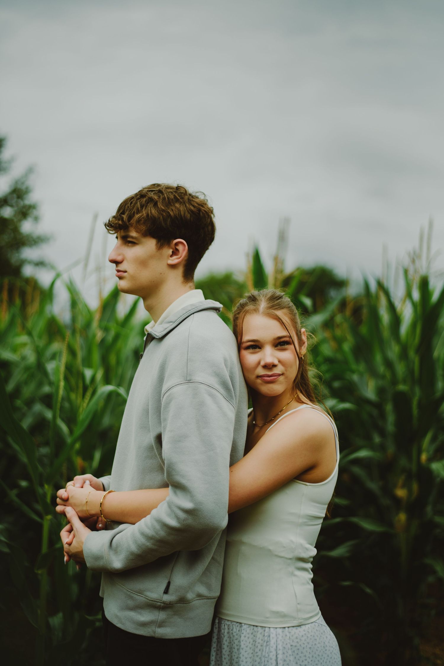 A young couple embraces in front of a tall cornfield on an overcast day.