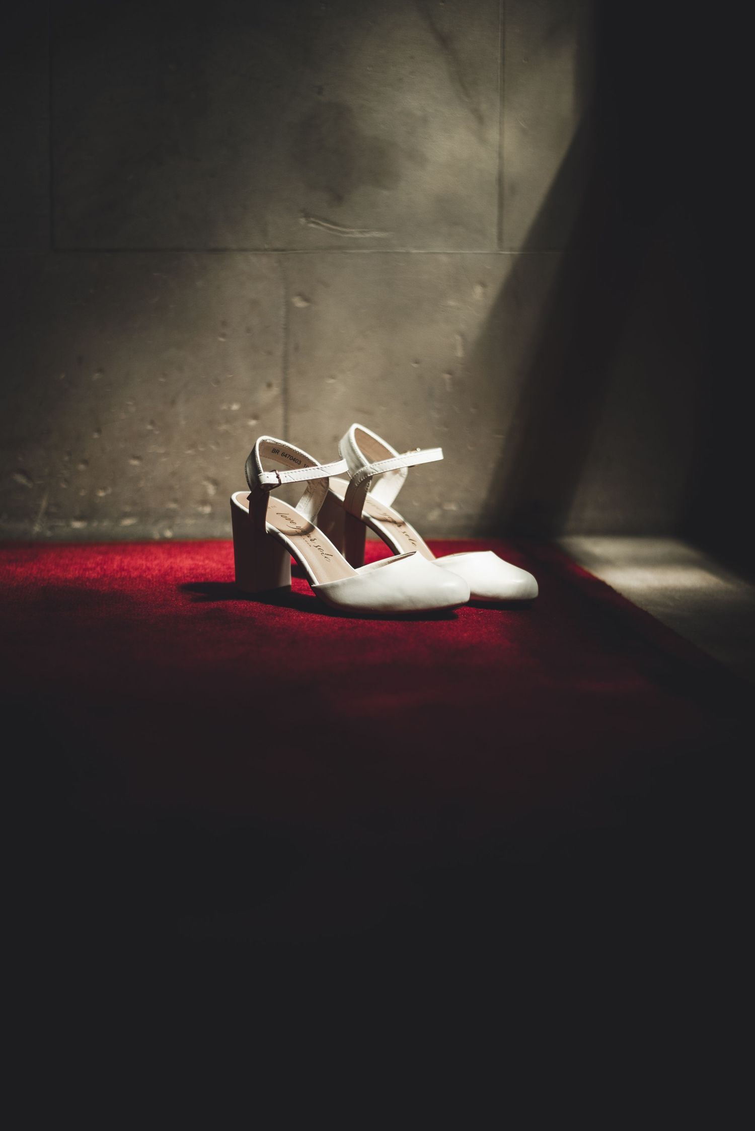 A pair of white vintage-style Mary Jane heels with ankle straps on a dark surface illuminated by dramatic red lighting.