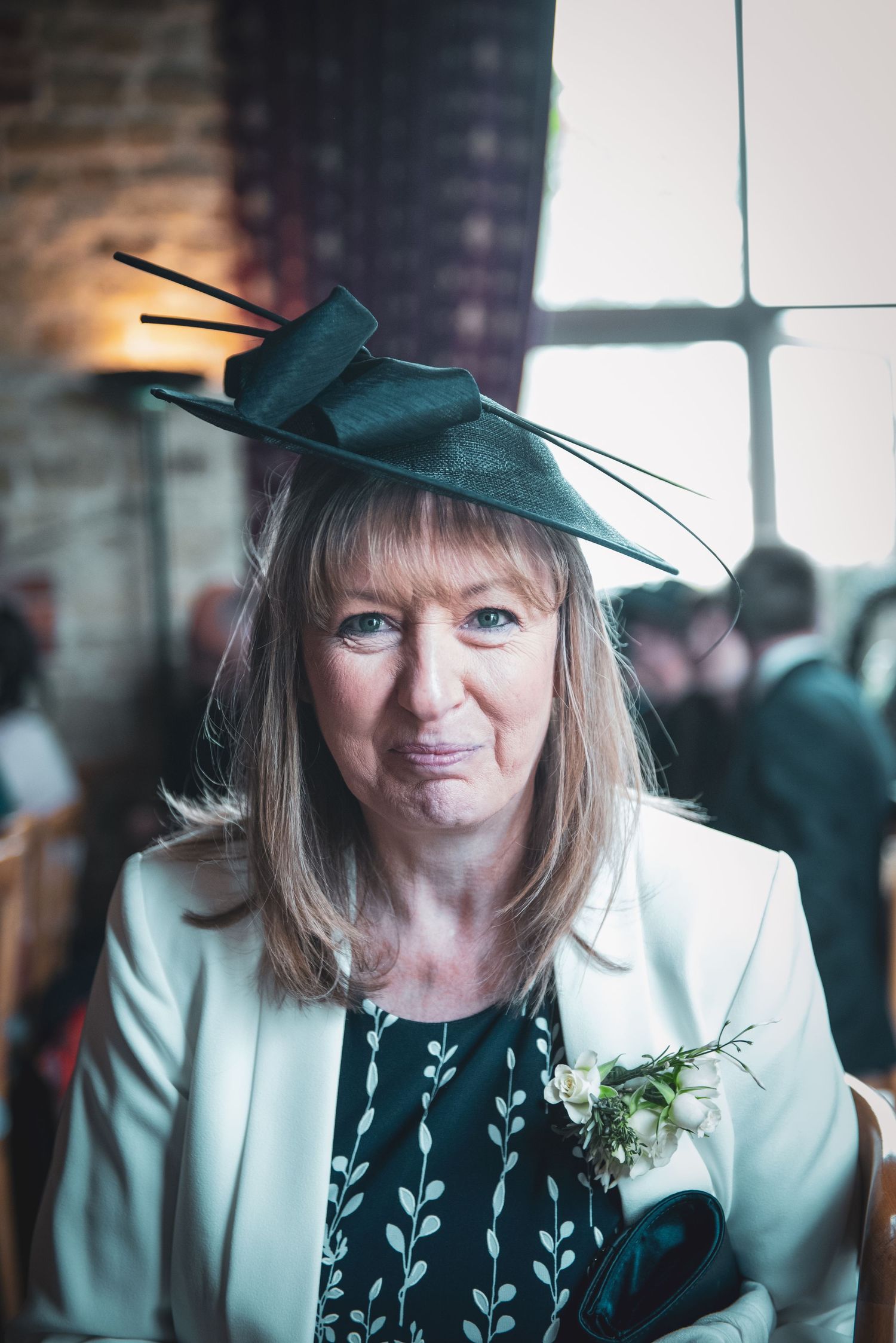 Wedding guest in teal hat and white blazer at indoor venue.