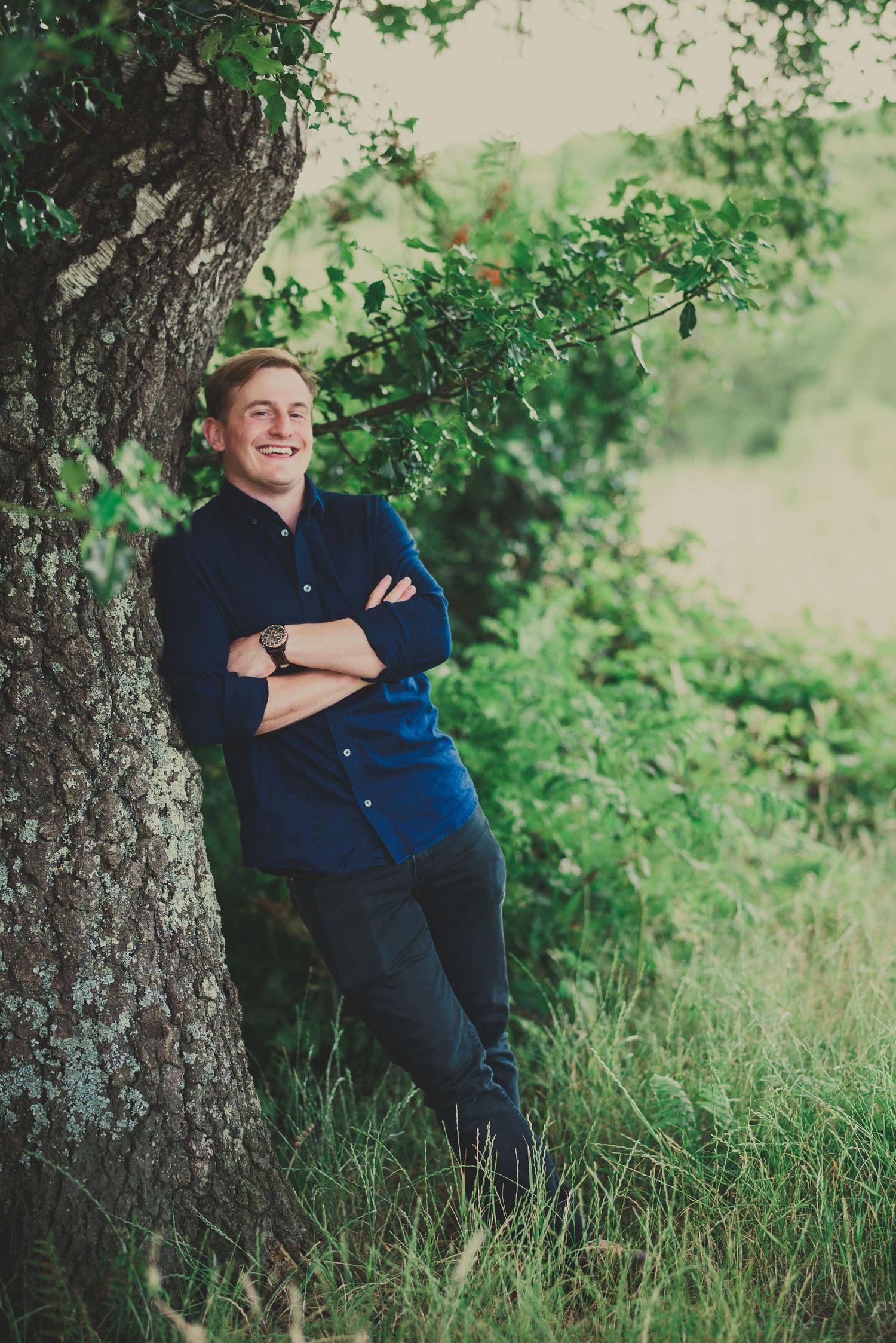 A casual portrait of someone leaning against a tree trunk in a navy blue shirt surrounded by green foliage.