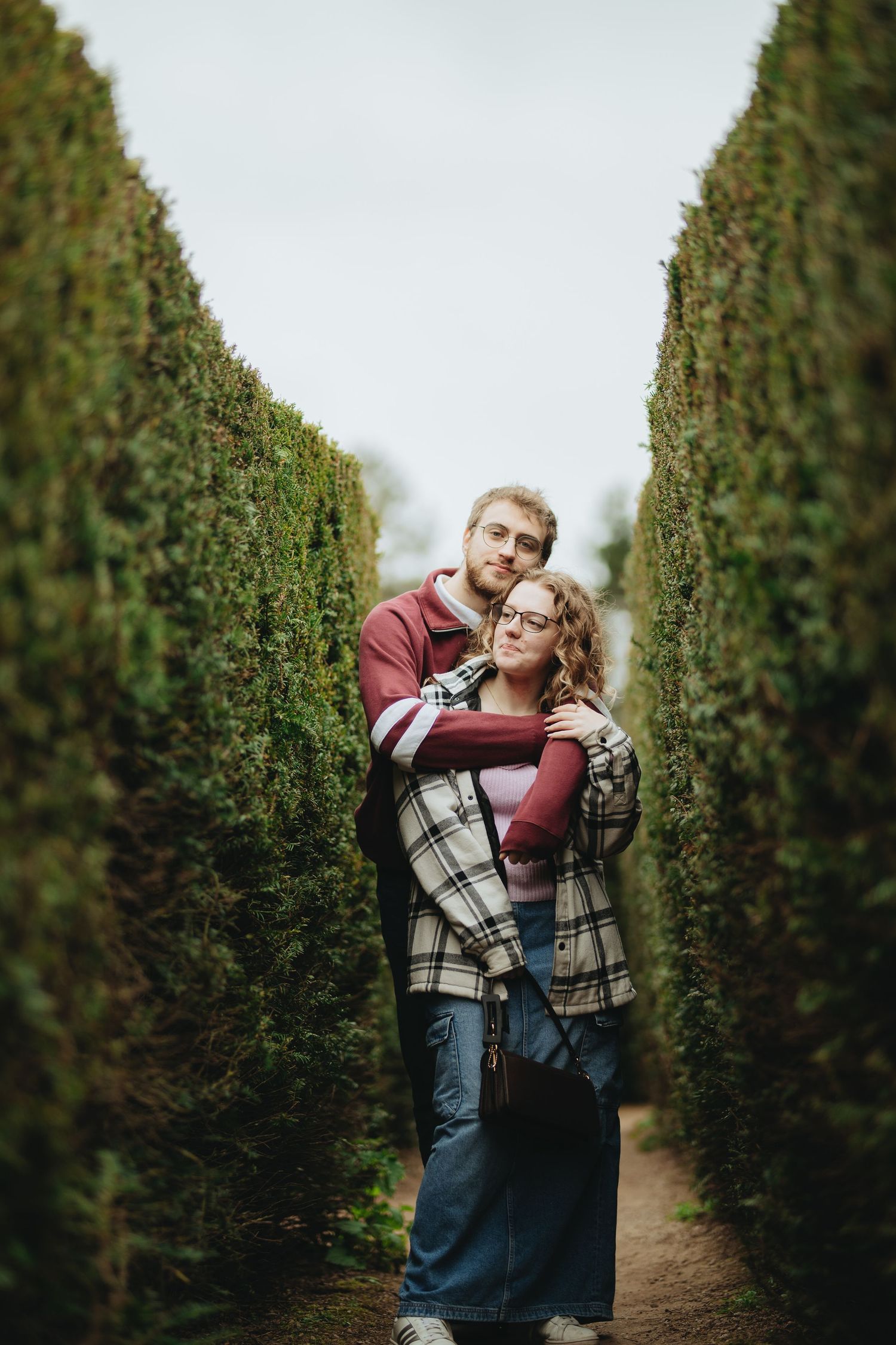 Couple embracing in a green hedge maze pathway, man hugging woman from behind, both smiling at camera.