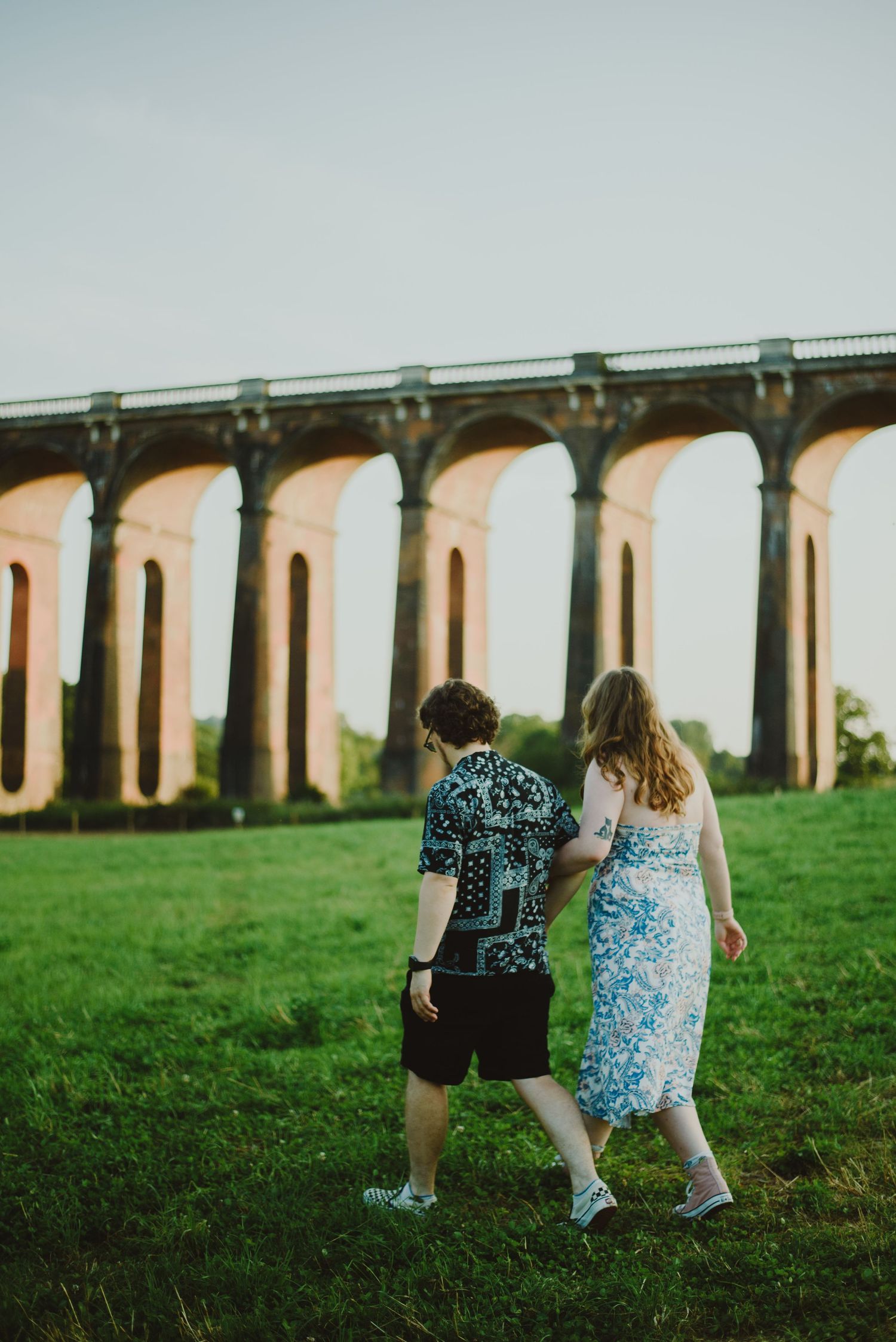 Two people in patterned clothing walk together on grass near a grand arched stone viaduct on a misty day.