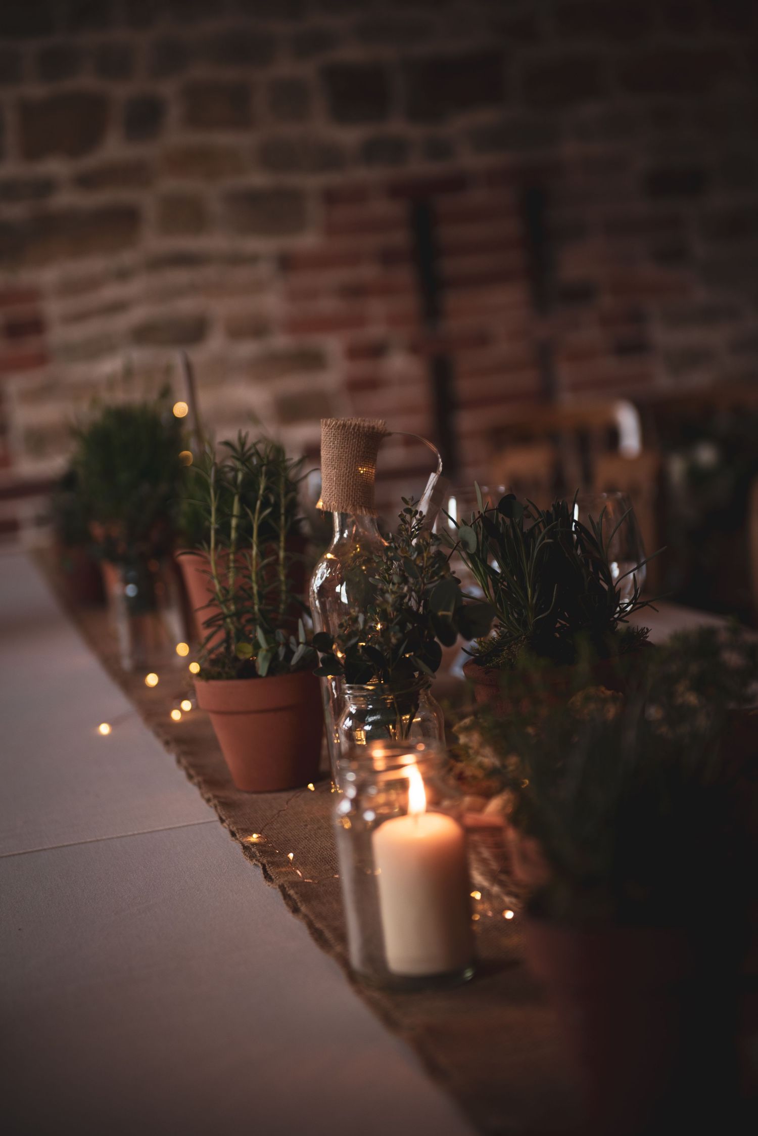 Romantic wedding table decor with potted herbs and glowing candles against brick wall.