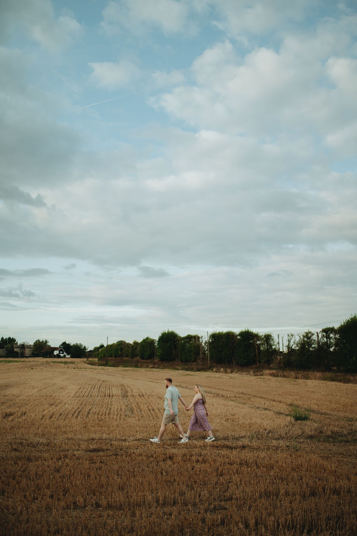 A couple walks hand in hand through a golden harvested field under a cloudy sky with trees in the background.