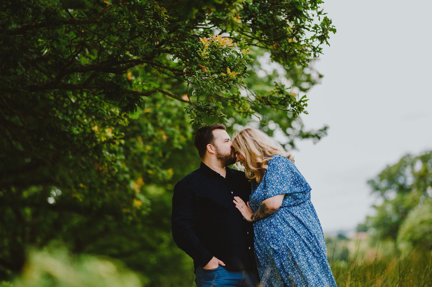 A couple embraces under green tree branches while wearing casual attire on an overcast day.