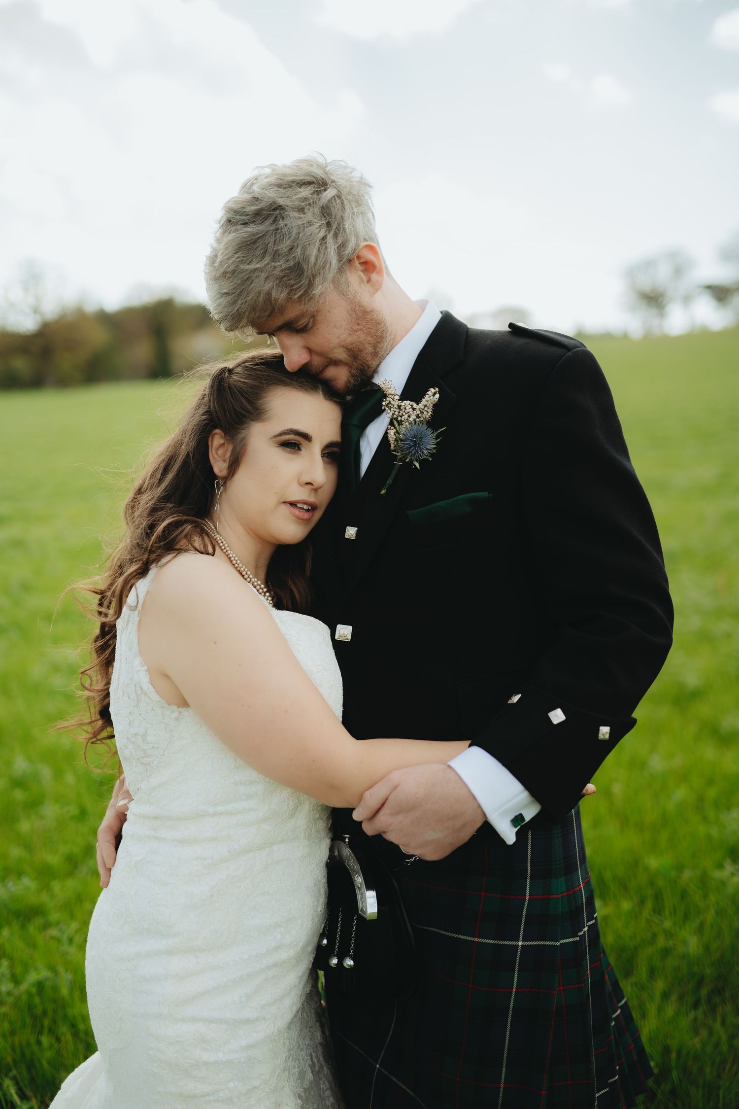 Bride in white gown and groom in Scottish tartan kilt embrace tenderly in a lush green field.