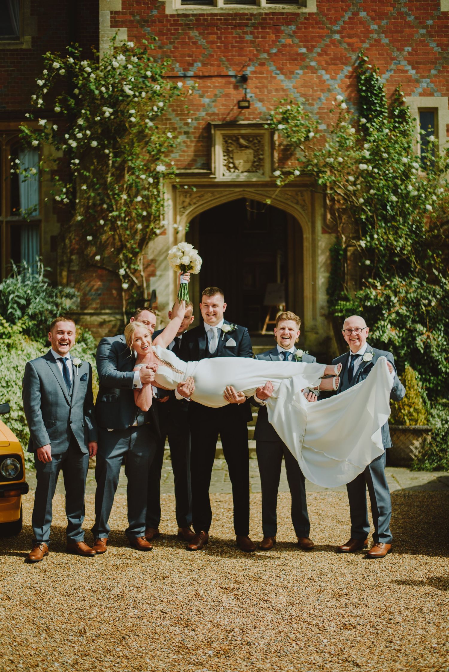 Wedding party stands in line holding up a flowing white dress train against red brick building exterior.