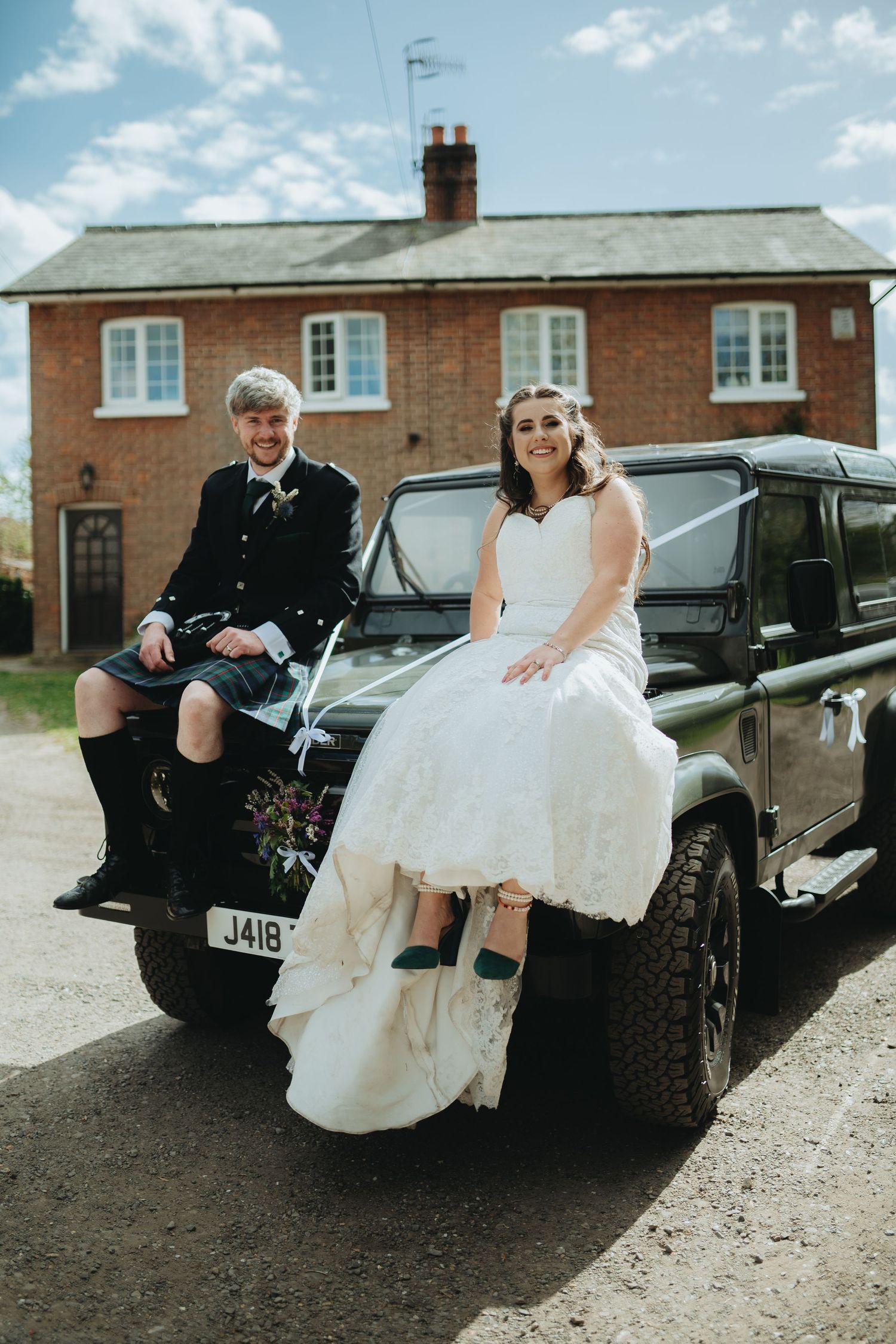 Bride in white gown and groom in Scottish kilt sit on a dark green Land Rover outside a brick house.
