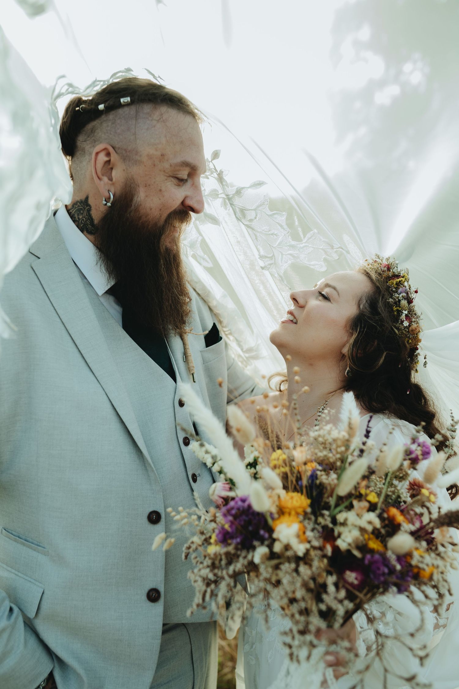 A bearded groom in a gray suit gazes at his bride holding a colorful wildflower bouquet at a boho wedding.