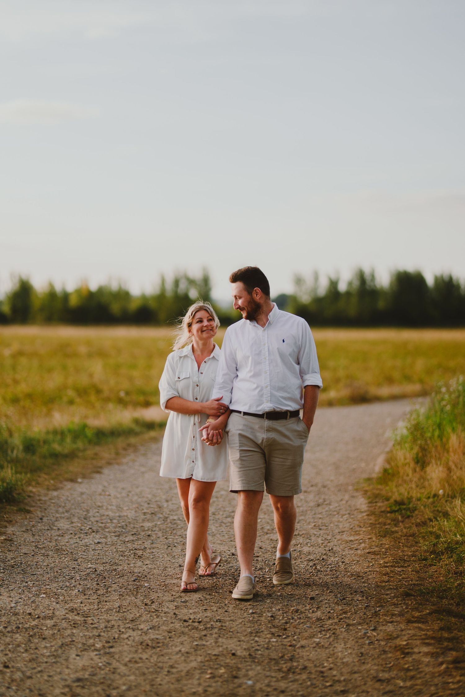 A couple walks together on a dirt path through a sunny countryside field during golden hour.
