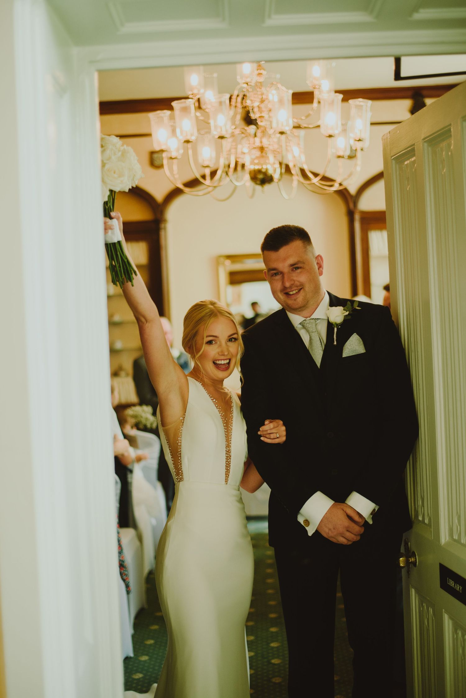 Joyful couple laughs together in a hallway during their wedding celebration with elegant chandeliers overhead.