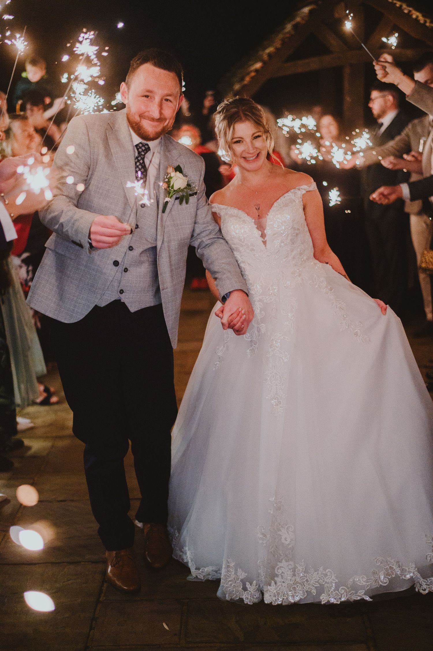 Newlyweds walk through sparkler exit at nighttime wedding celebration with glowing lights and rustic barn backdrop.