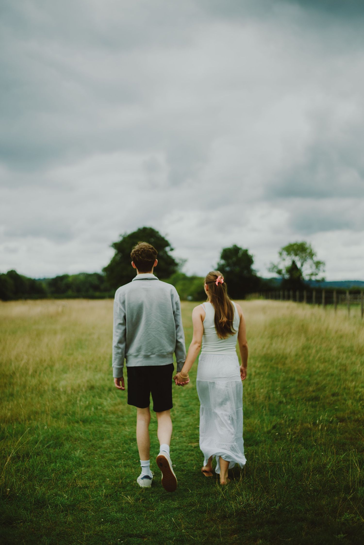 A couple walks hand in hand through a serene meadow on a cloudy day, with trees visible in the distance.