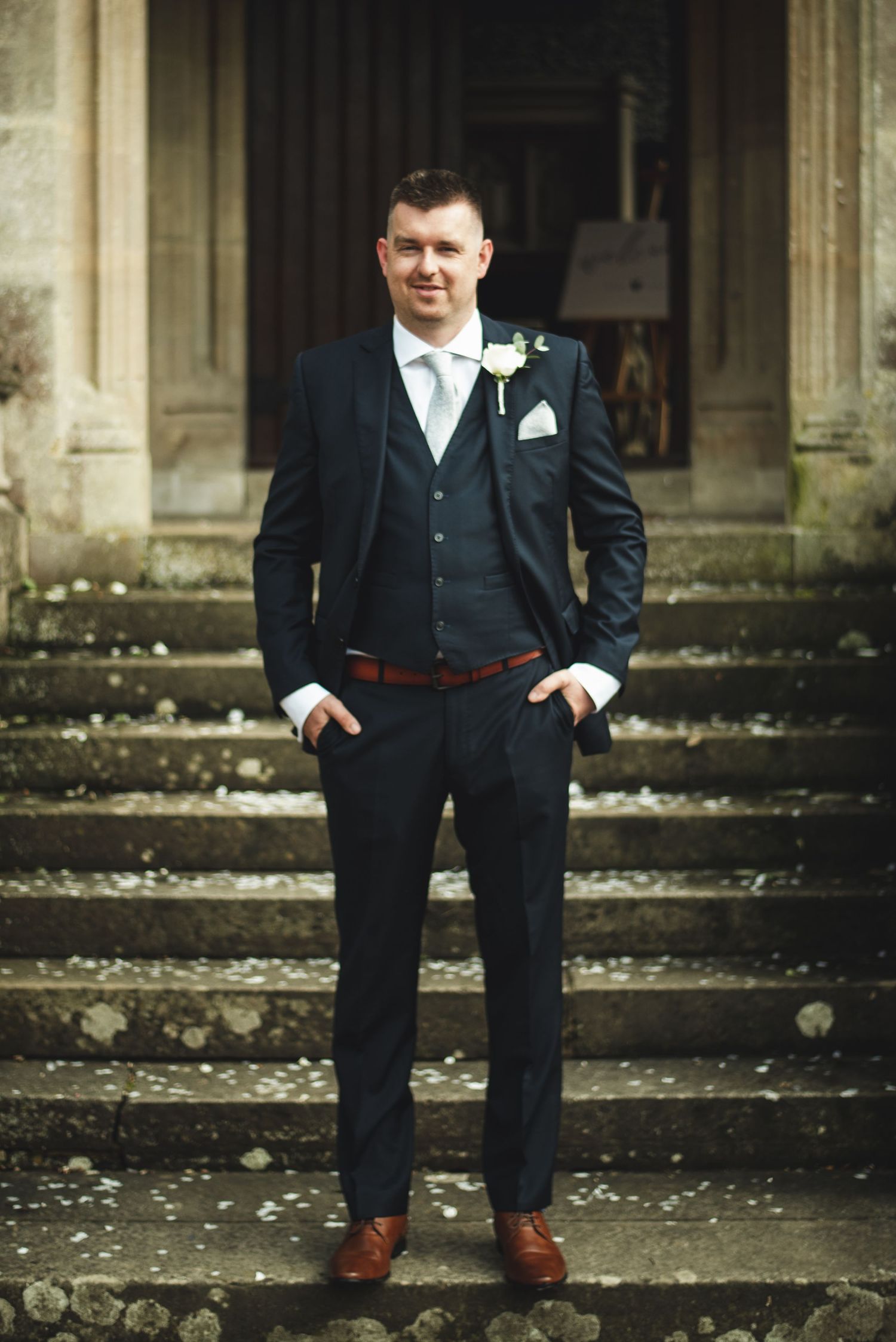 Groom in navy three-piece suit stands confidently on stone steps of historic building.