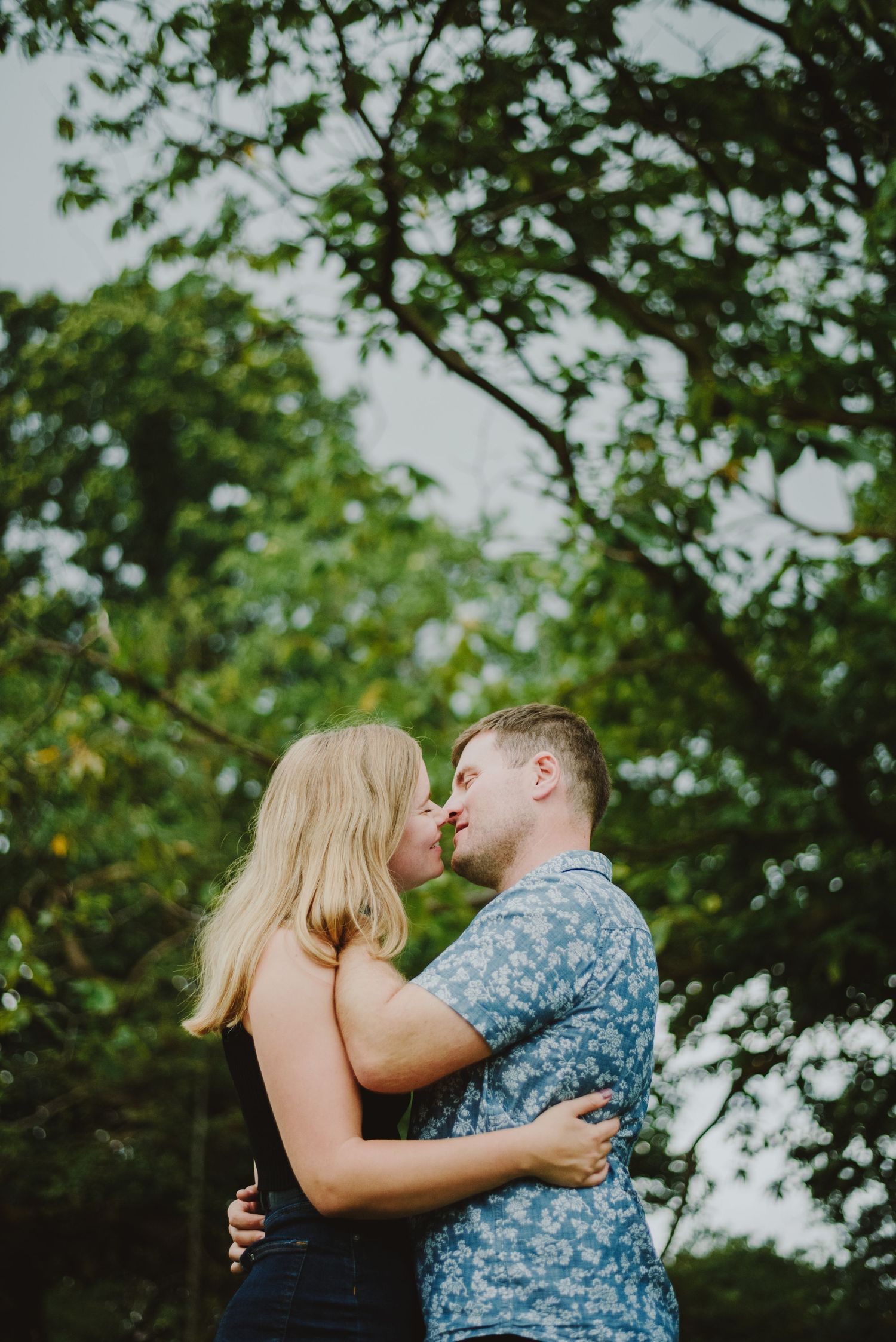 A romantic couple embraces outdoors under green trees on a summer day.