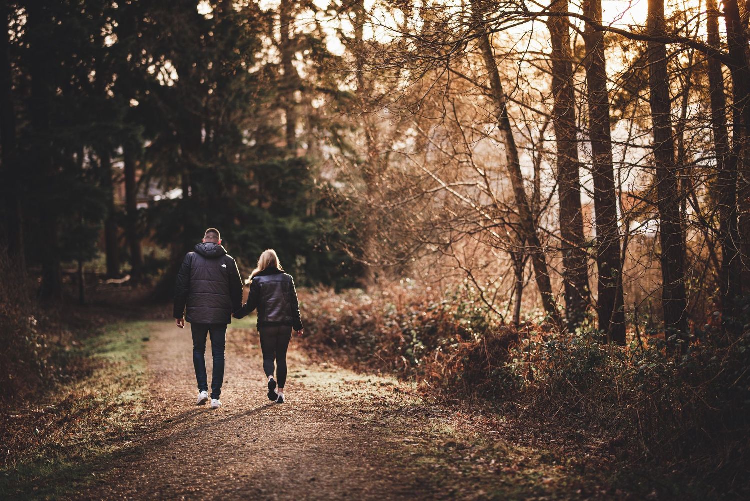 Kent engagement shoot.Two figures walking along a sunlit forest path during golden hour.