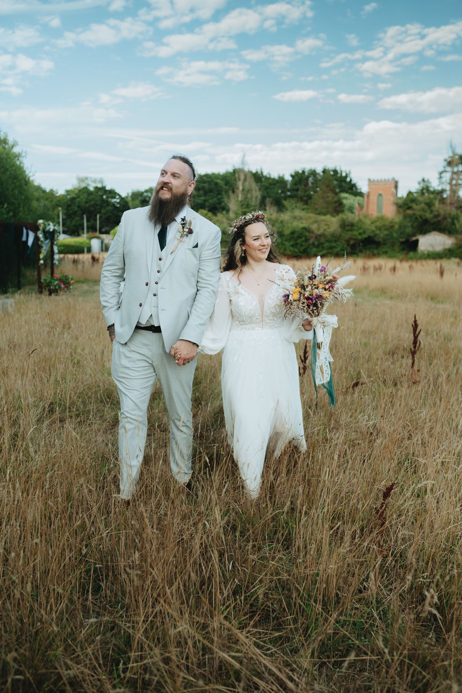 Bride and groom holding hands walking through a golden field, bride in white lace gown holding colorful bouquet, groom in gray suit.