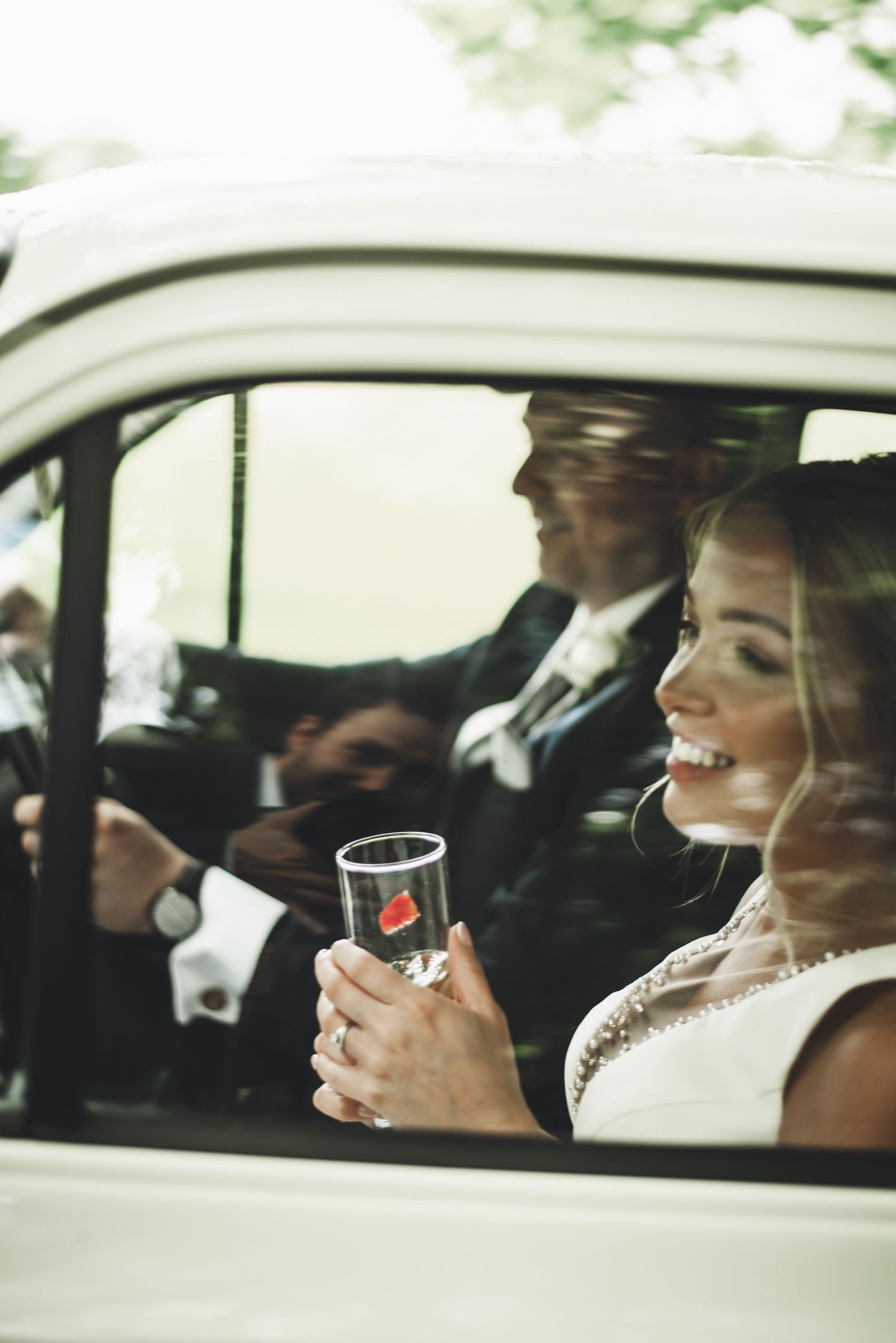 Wedding party members smile and laugh inside a car during celebrations.