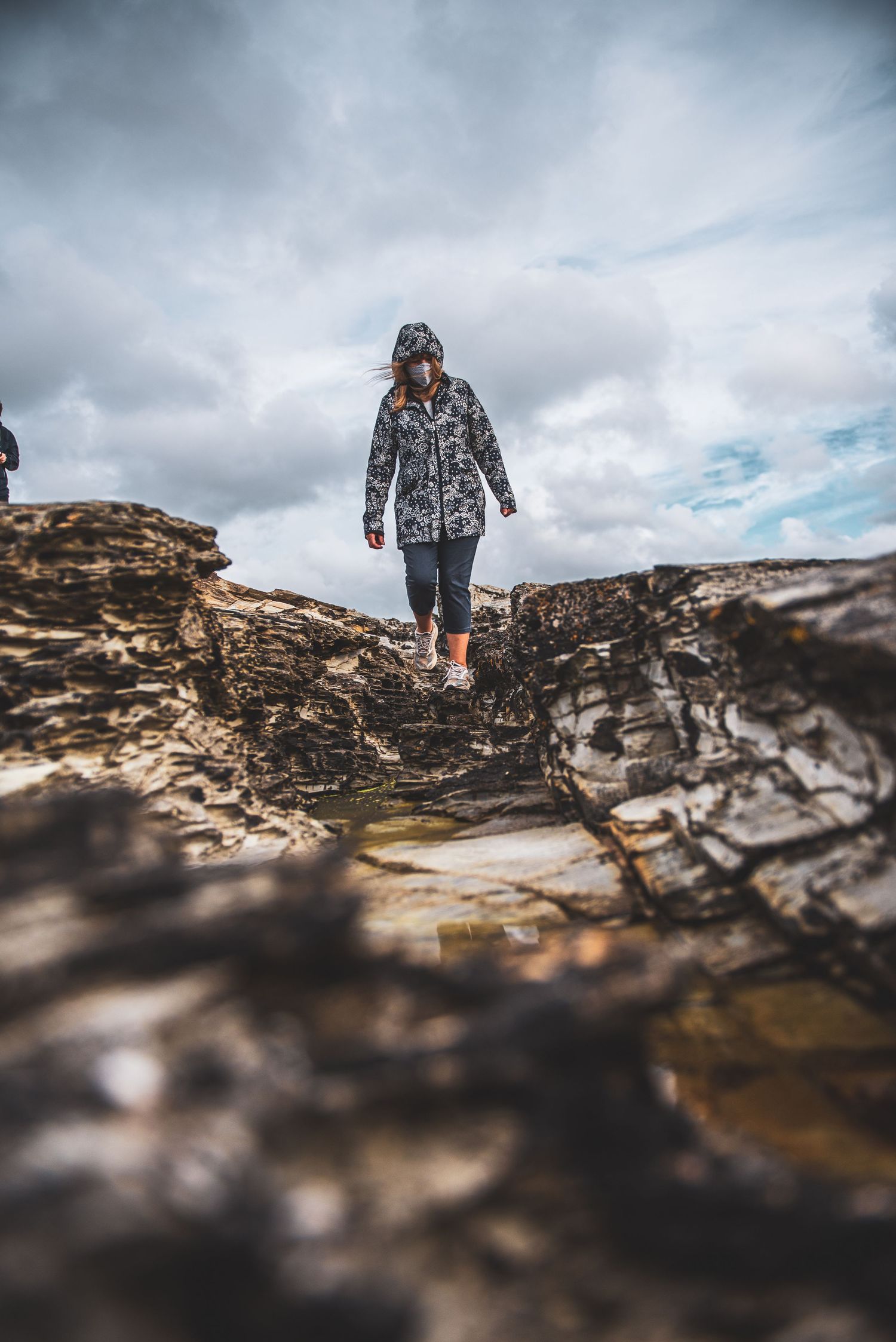 A hiker wearing a patterned jacket walks across rocky terrain under dramatic cloudy skies.
