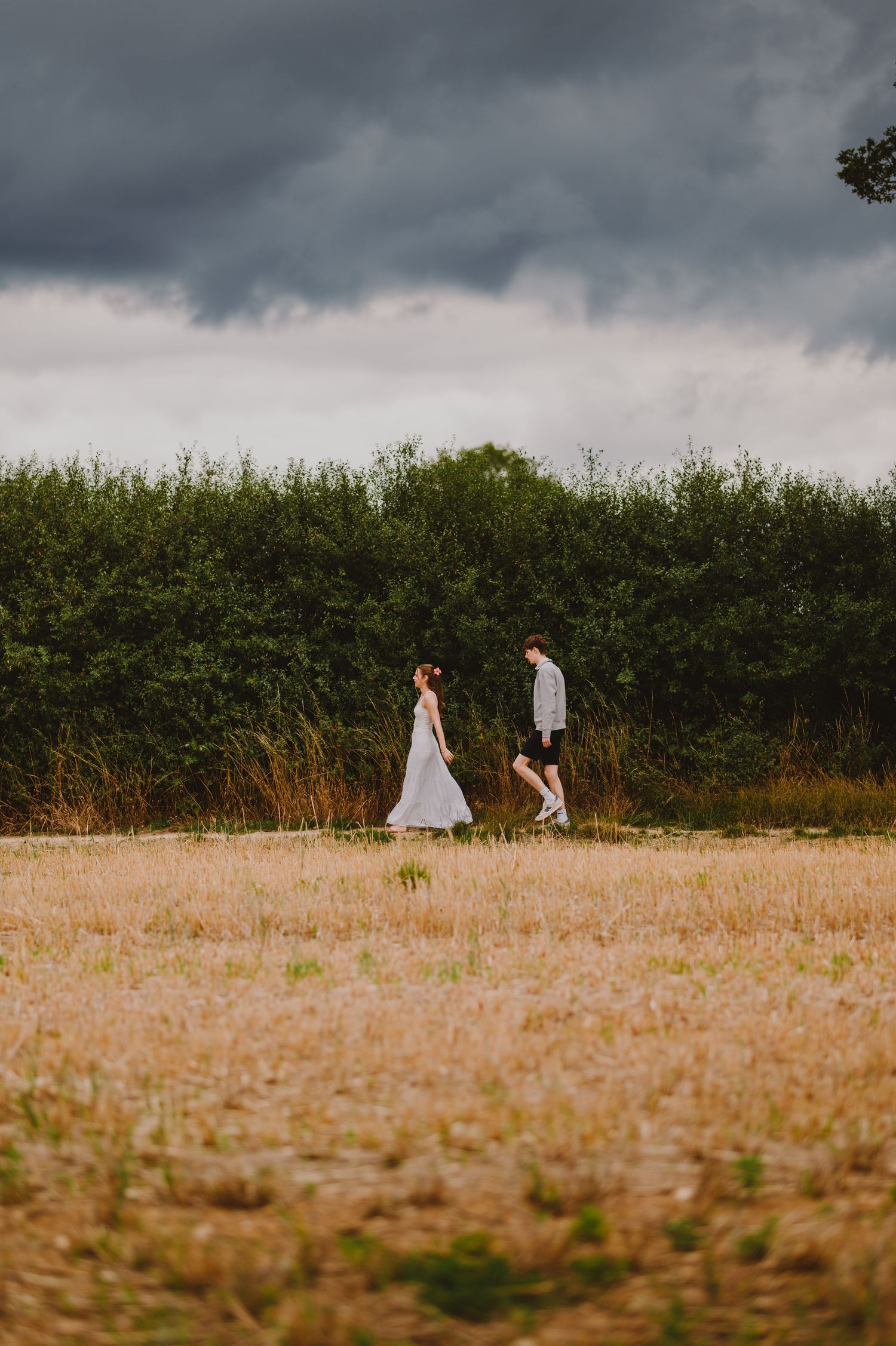 Two people walk along a grassy path under moody storm clouds during a countryside photo session.