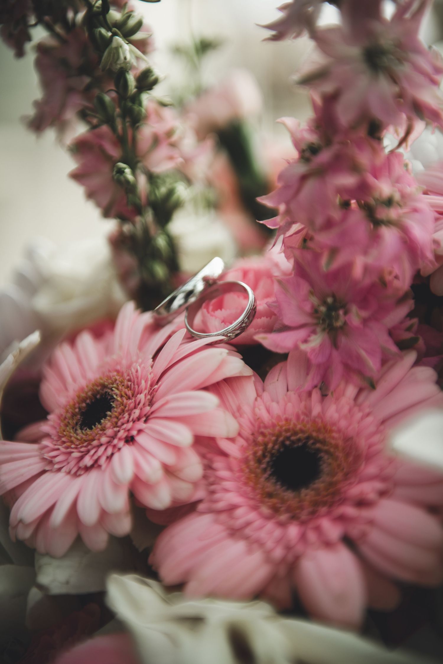 A wedding ring rests atop pink gerbera daisies and delicate flowers in a romantic floral arrangement.