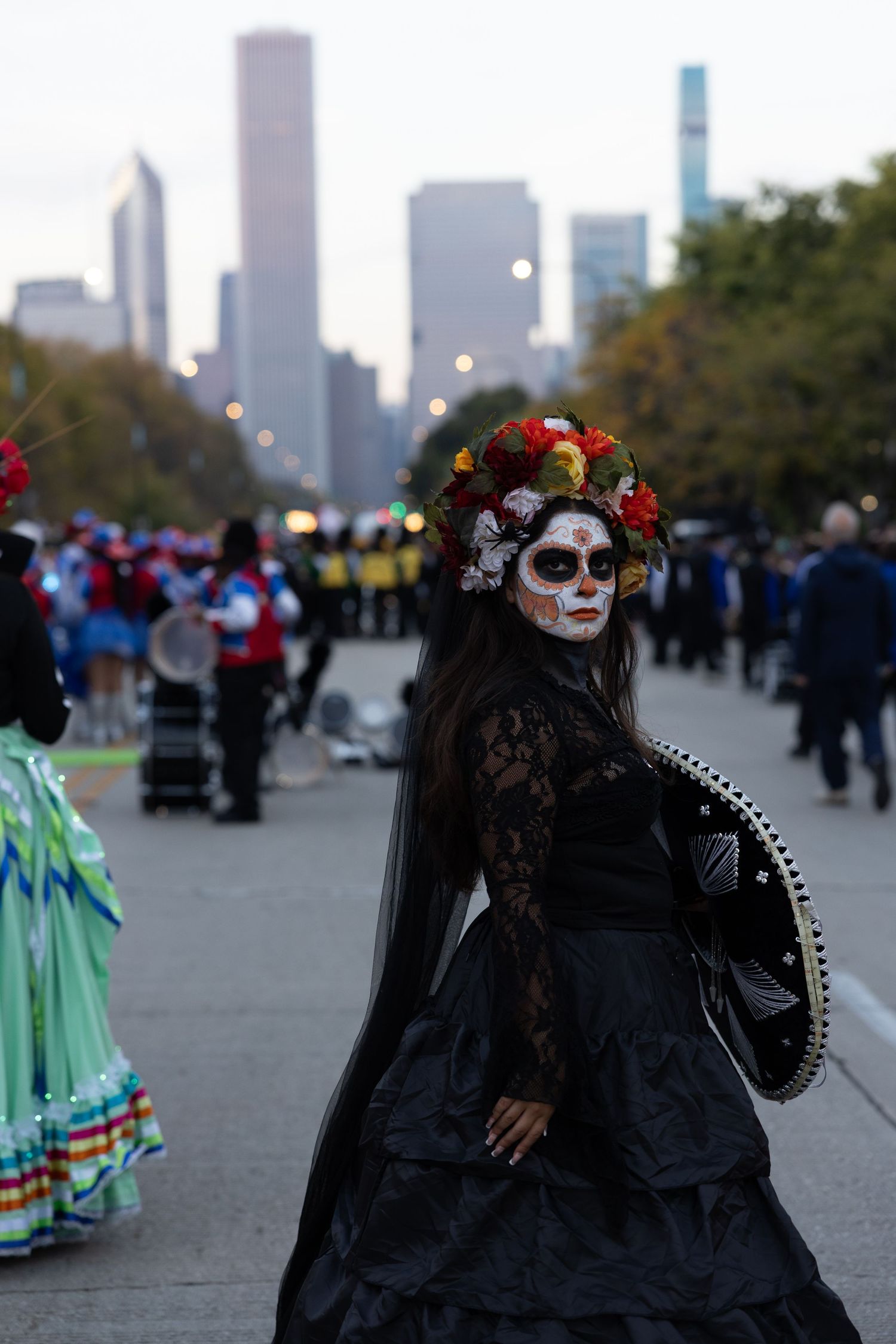 Woman in black lace dress with Catrina skull face paint and floral crown stands in a Day of the Dead parade with city skyline behind.