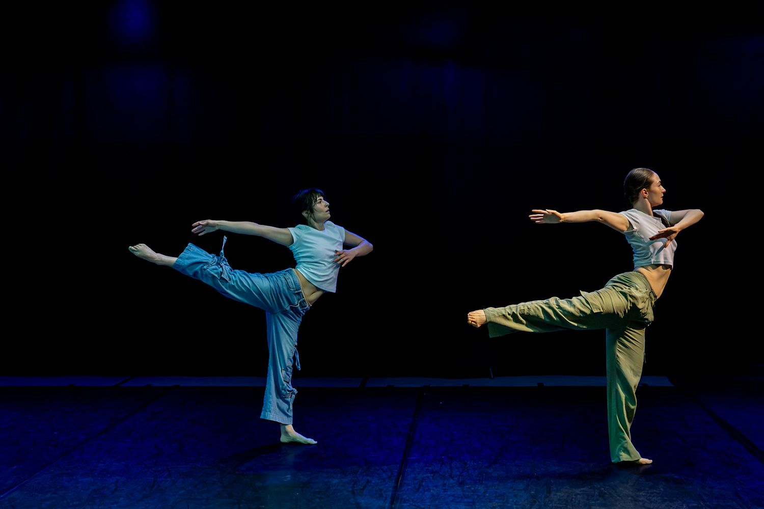 Two contemporary dancers in light blue and green outfits perform synchronized leg extensions on a dark stage with blue lighting.