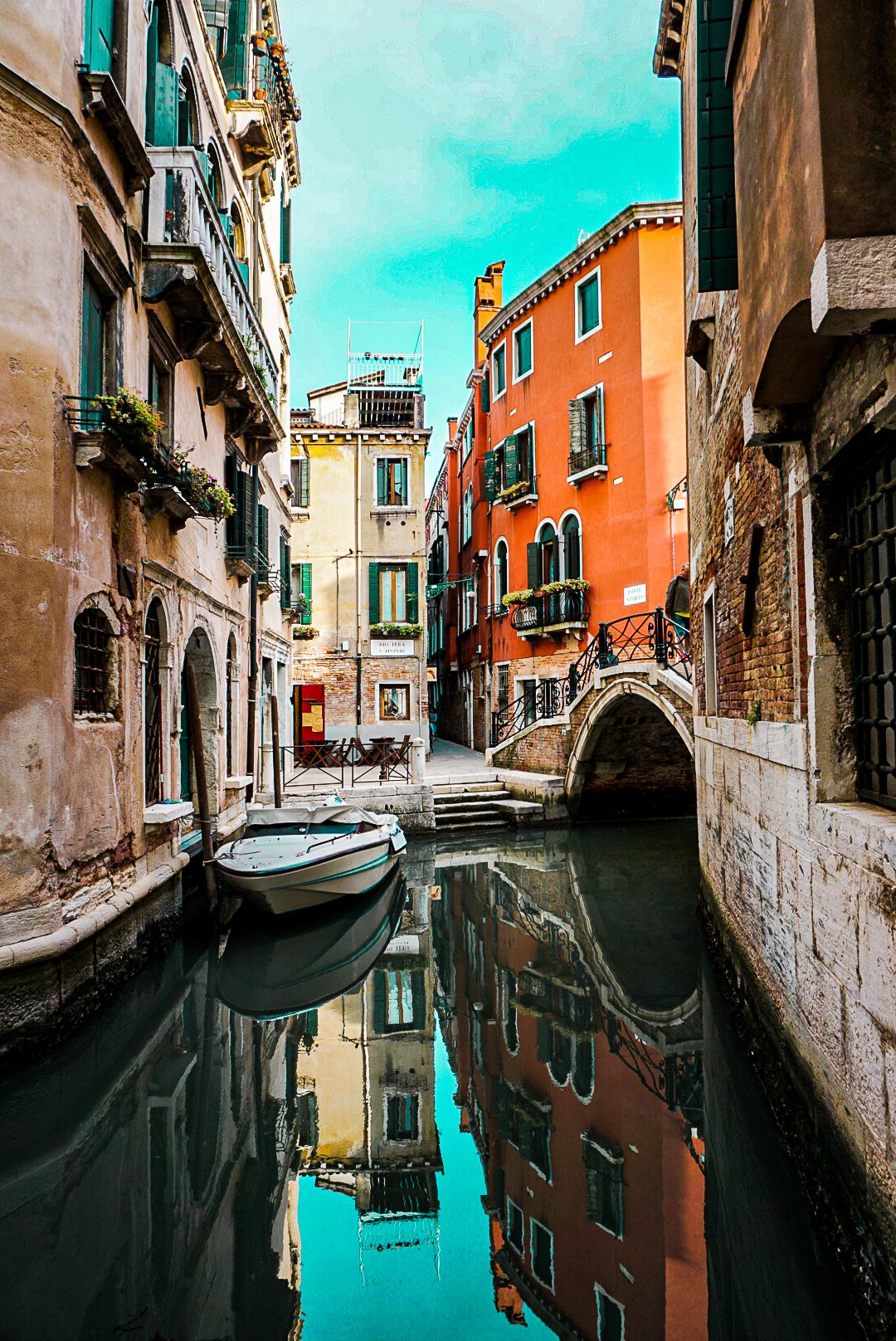 A serene Venice canal reflects colorful historic buildings and a small white boat beside a stone bridge.