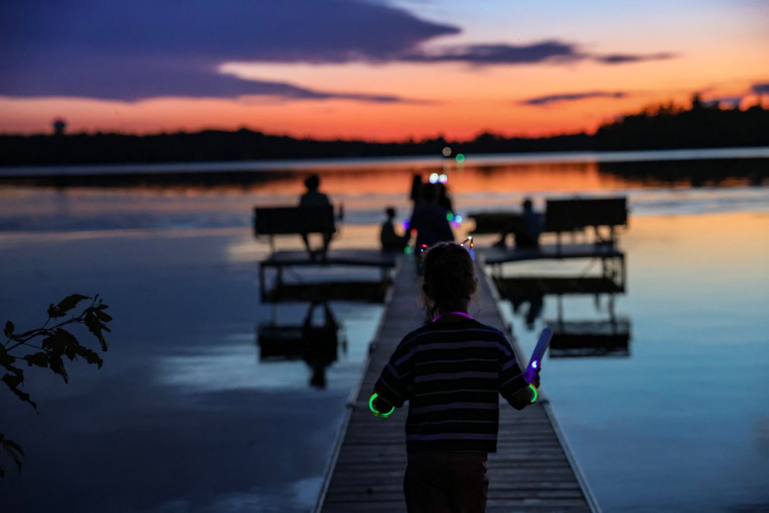 Child holding glow sticks on a wooden dock at sunset with family silhouettes gathered near the calm lake.