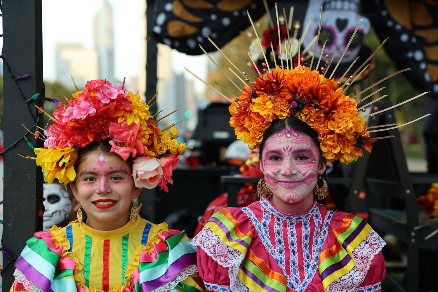 Two women in colorful embroidered dresses and floral headdresses with painted faces celebrate Day of the Dead in Mexico.