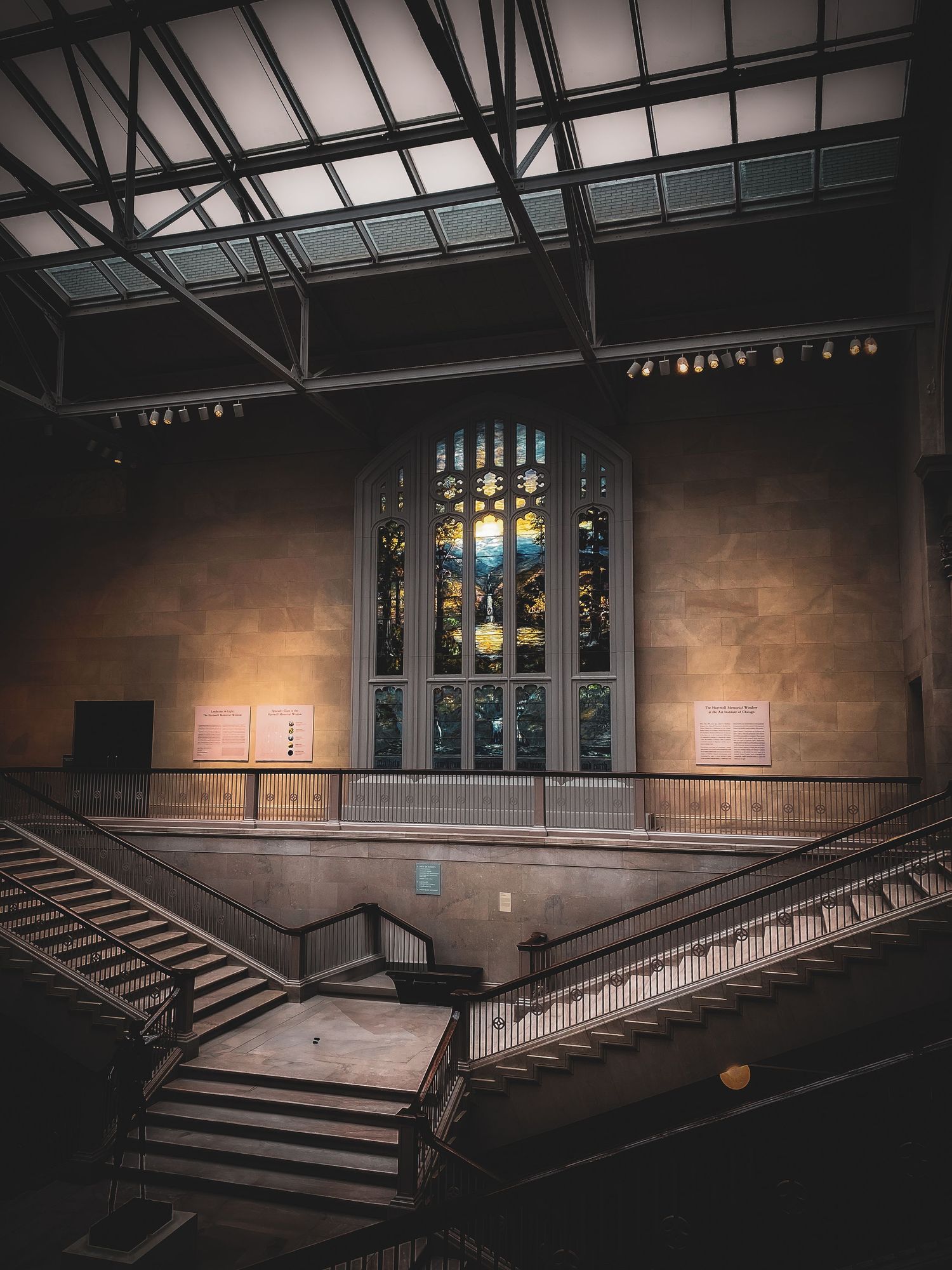 A grand museum interior features a stunning stained glass window above symmetrical staircases in dim light.
