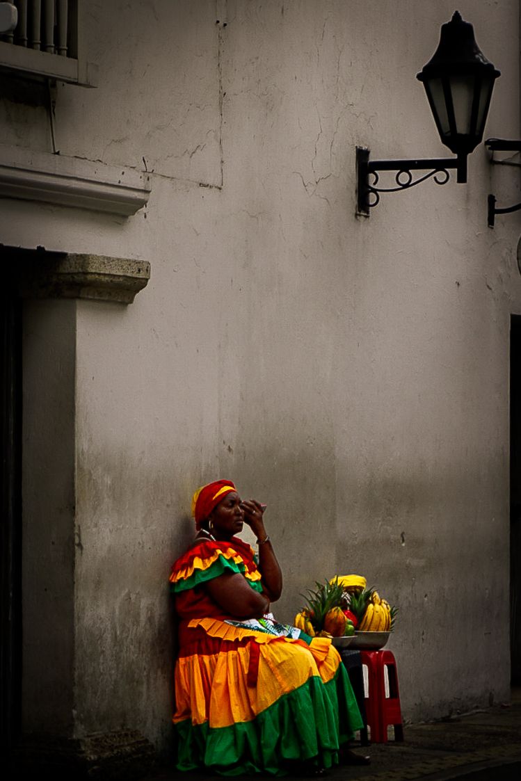 A woman in vibrant traditional Colombian dress sells tropical fruit beside a white wall and street lamp.