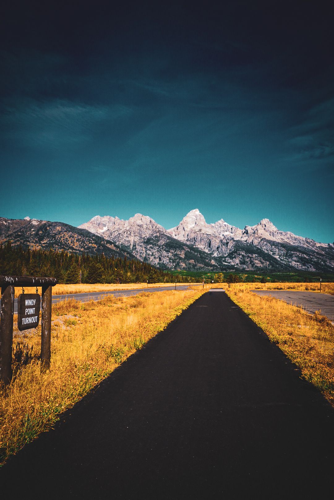 Narrow pathway leading toward snow-capped Grand Teton peaks with golden meadows and dark dramatic sky in Wyoming.