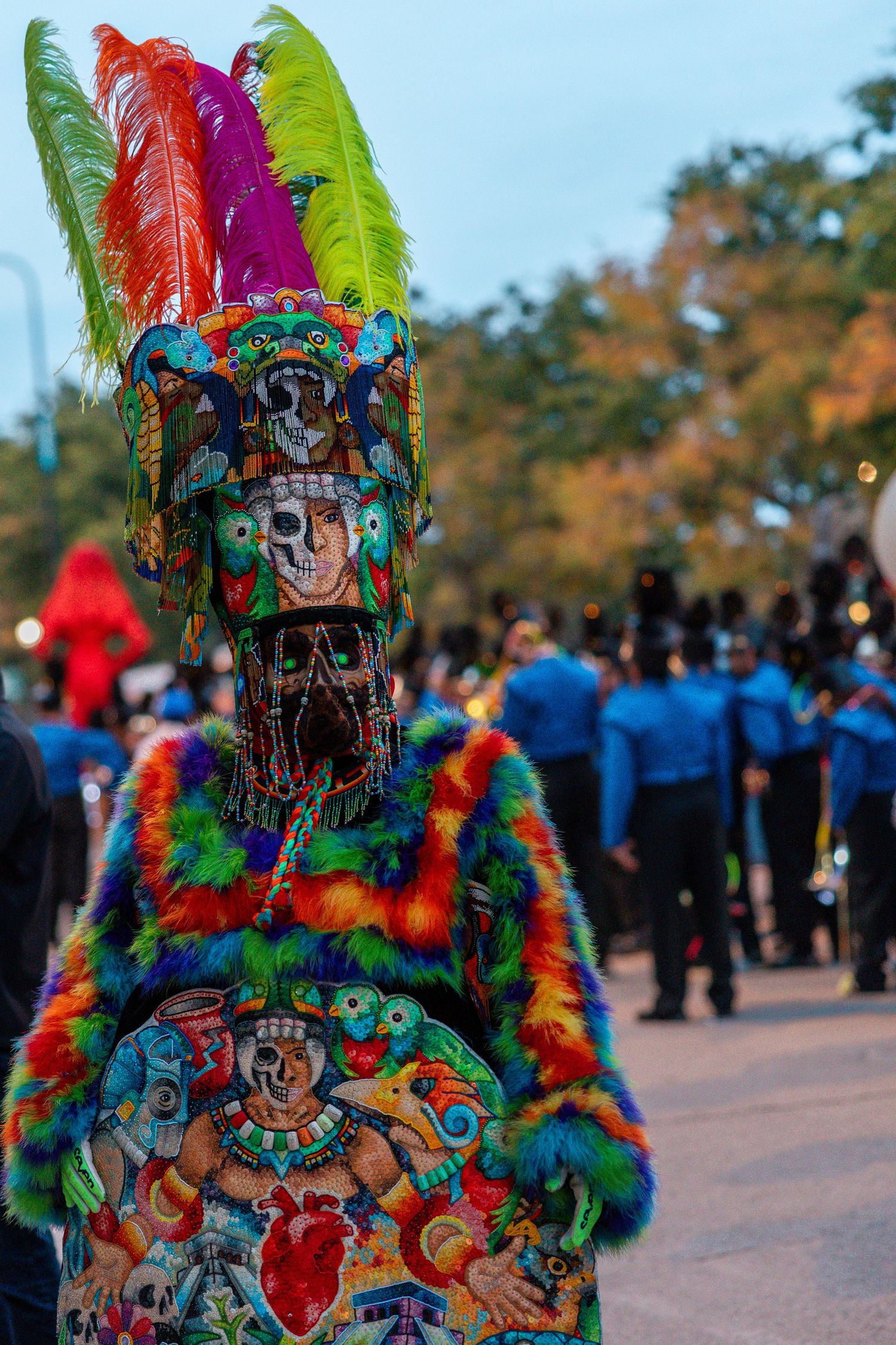 Parade performer in a colorful skull mask, feathered headdress and embroidered Day of the Dead costume with a marching band behind.