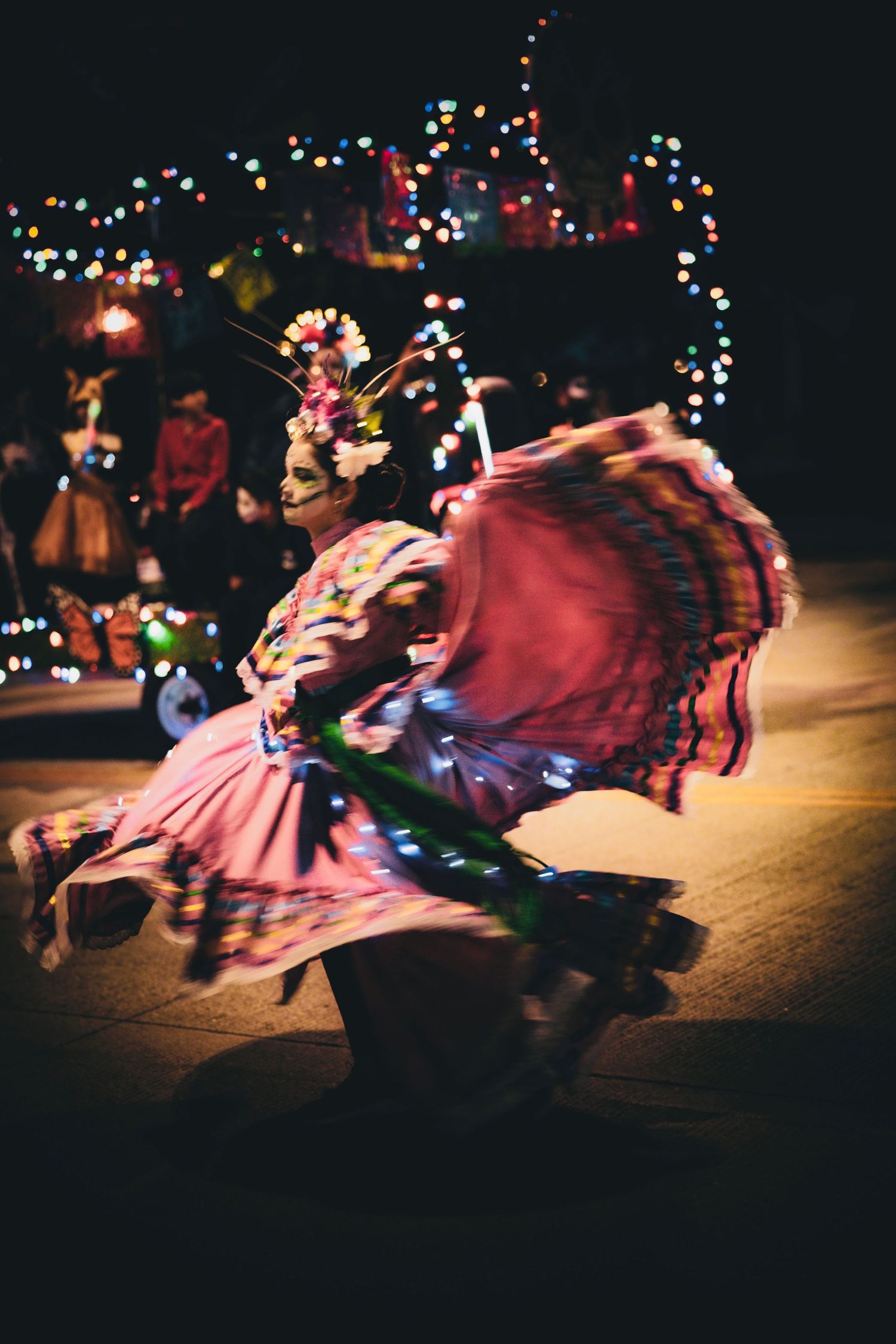 A folklórico dancer in a flowing pink ruffled skirt and floral headpiece twirls at night in front of festive string lights.