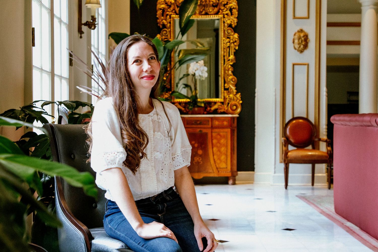 Smiling young woman in white blouse sitting in an elegant ornate interior with marble floors.