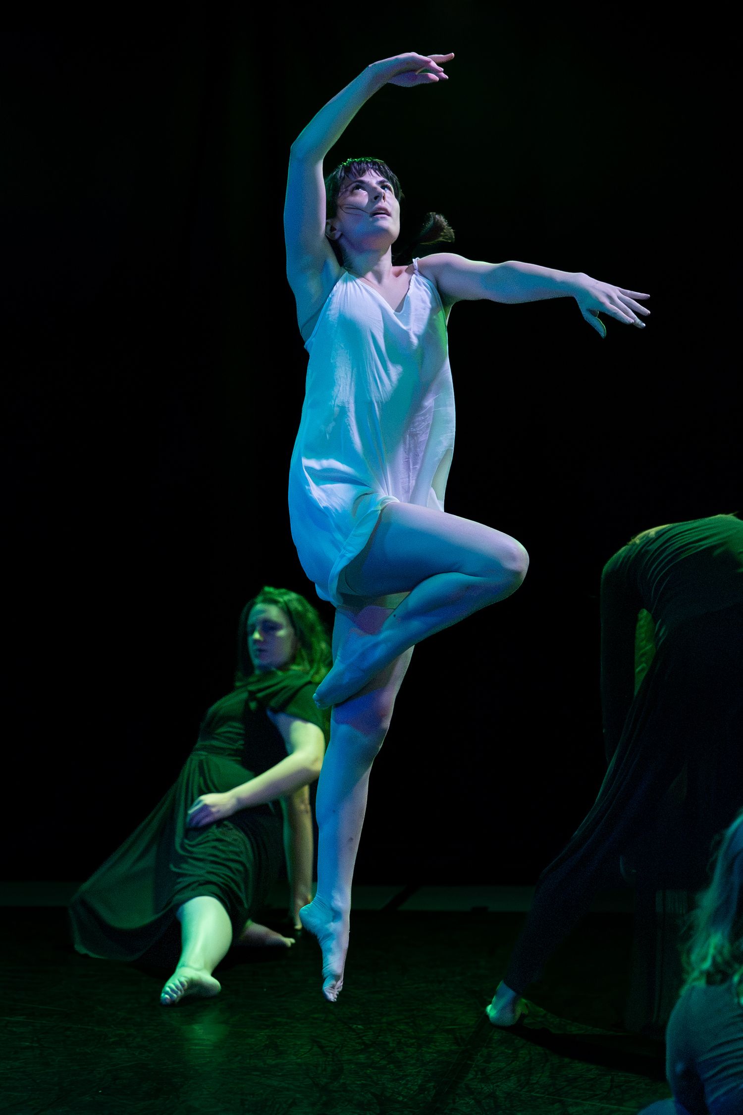 A dancer in a flowing white dress leaps mid-air with outstretched arms on a dark stage while another performer in green reclines nearby.