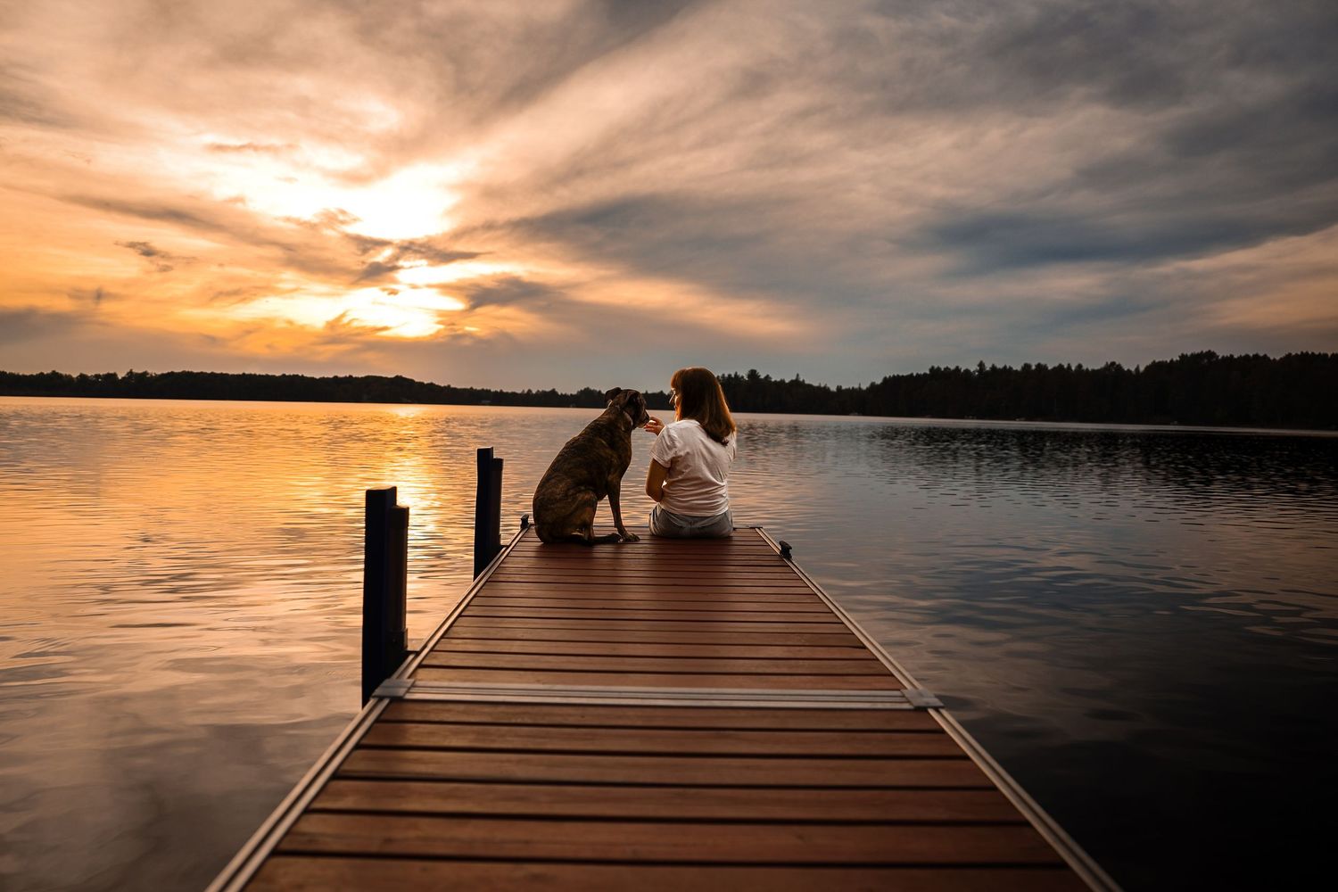 Woman and dog sit together on a wooden dock overlooking a calm lake at sunset under a dramatic golden cloudy sky.