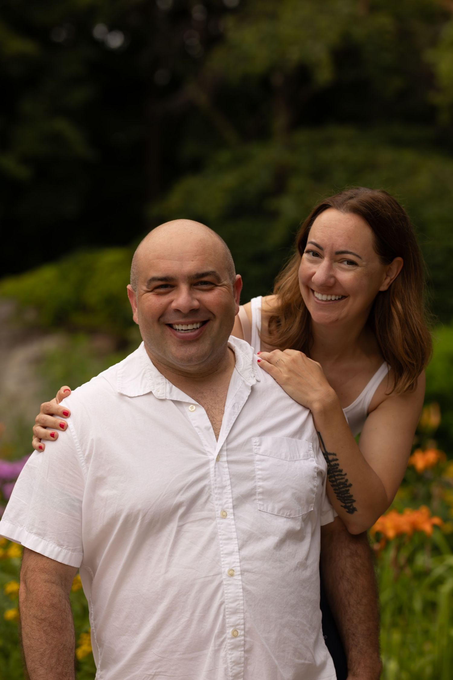Smiling bald man in a white button-up shirt posing with a smiling woman with long brown hair outdoors near orange flowers and greenery.