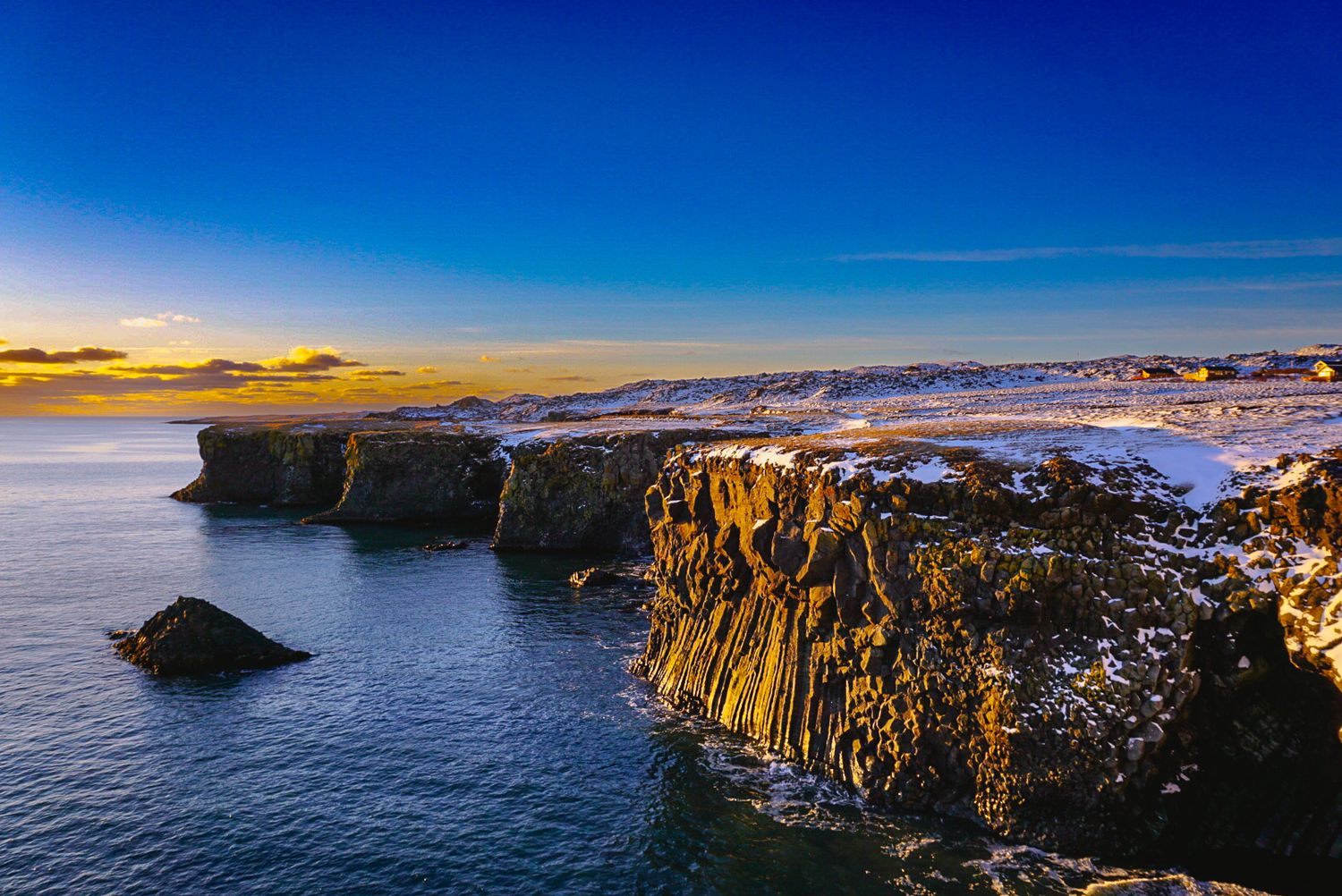 Dramatic coastal cliffs dusted with snow at sunset, with golden light reflecting off dark ocean waters under a vivid blue sky.