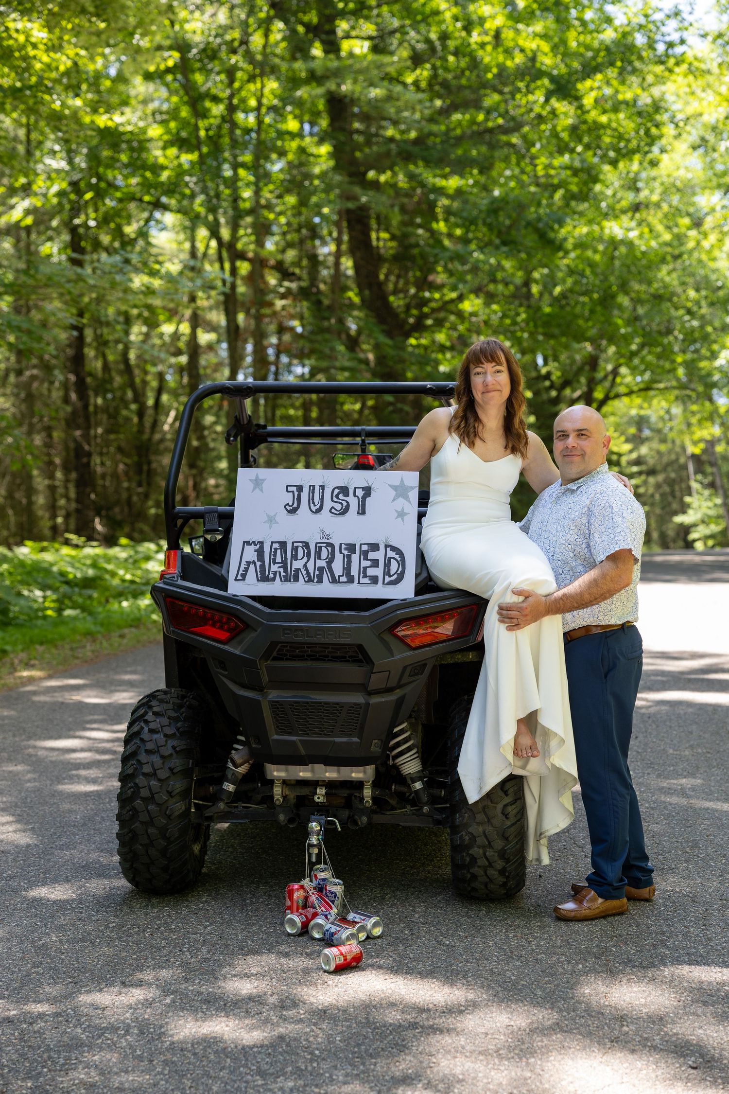 Newlywed couple posing beside an ATV with a Just Married sign in a lush green forest setting.