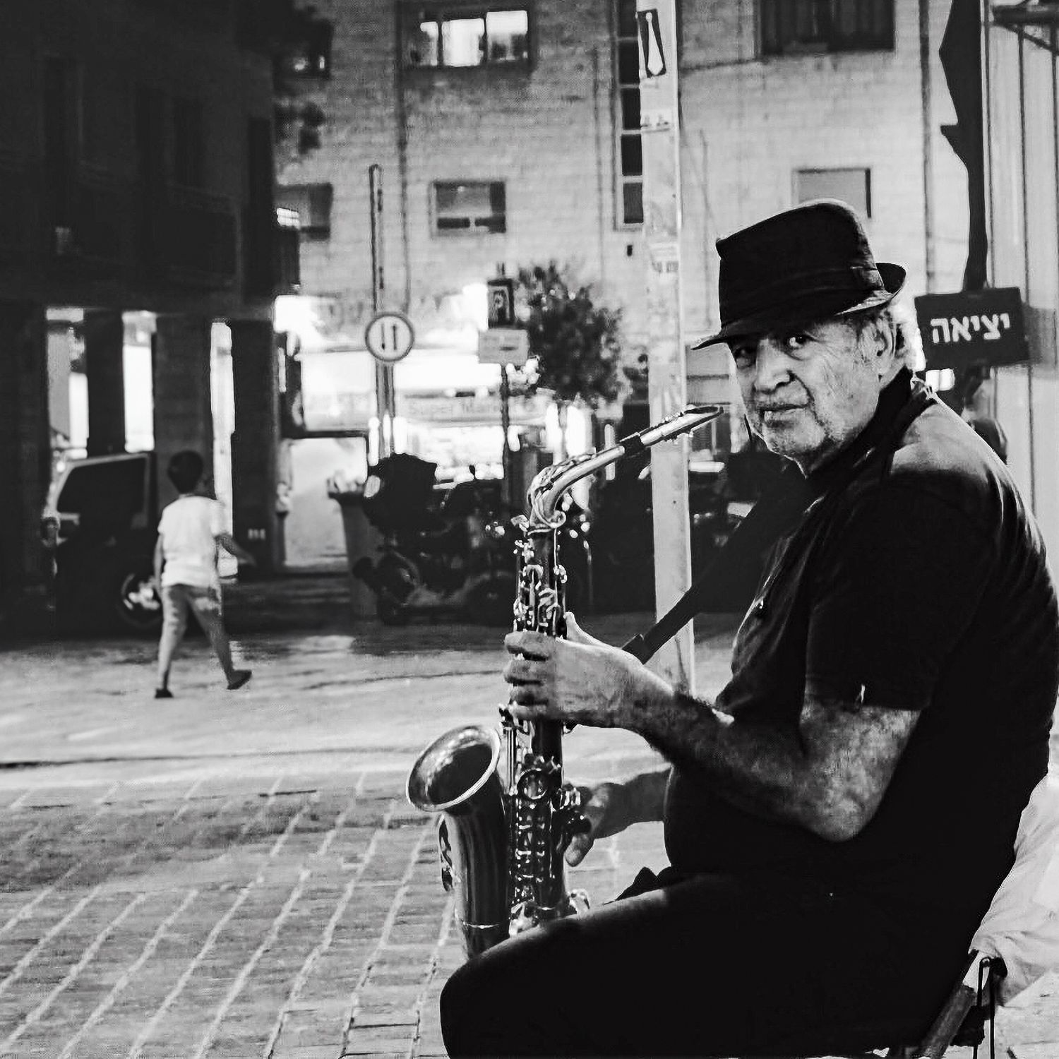 Black and white street photo of an elderly tattooed saxophonist performing on a city sidewalk at night.