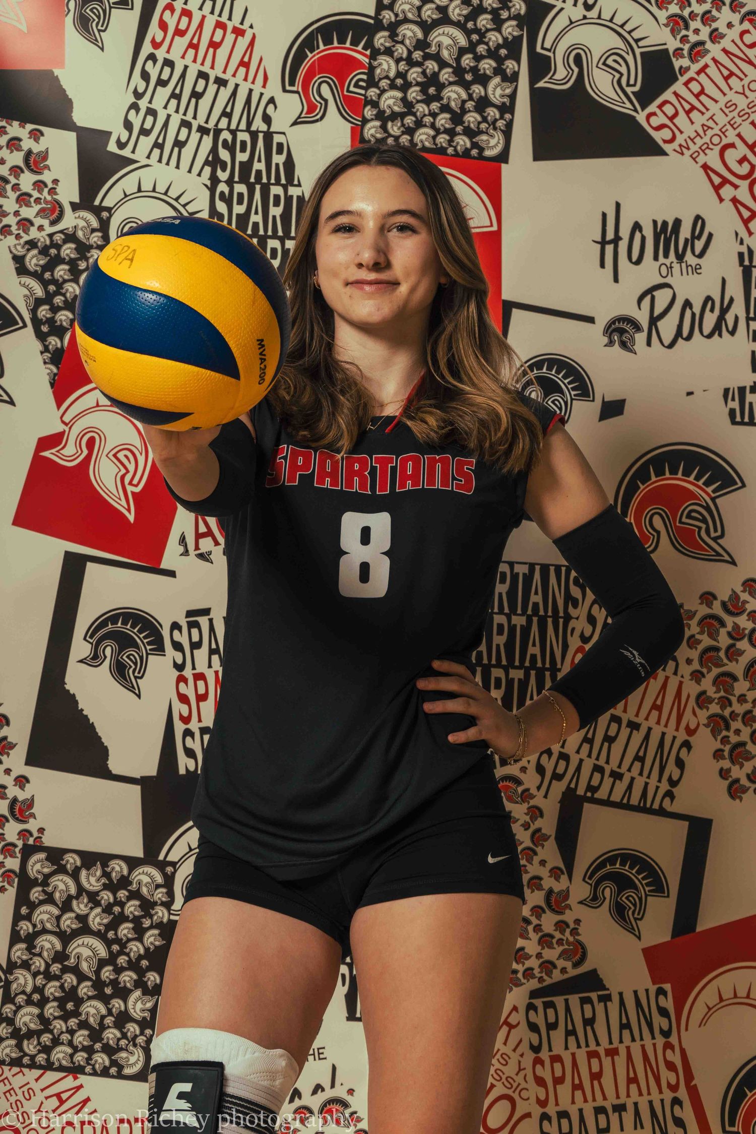 Athletes in black uniforms hold volleyballs in front of a decorative team logo wall.