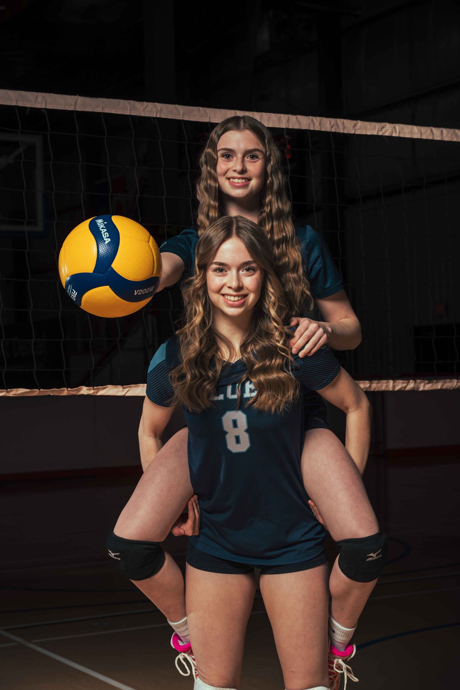 Volleyball teammates in navy uniforms pose together with volleyball on indoor court.