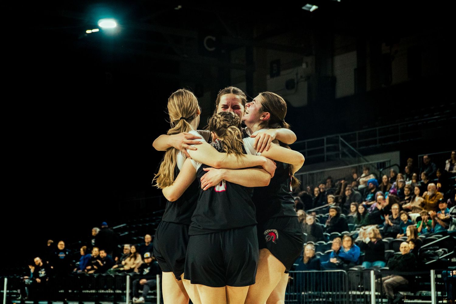 Athletes in black uniforms embrace in a group hug during an indoor Basketball game as spectators watch from the stands.